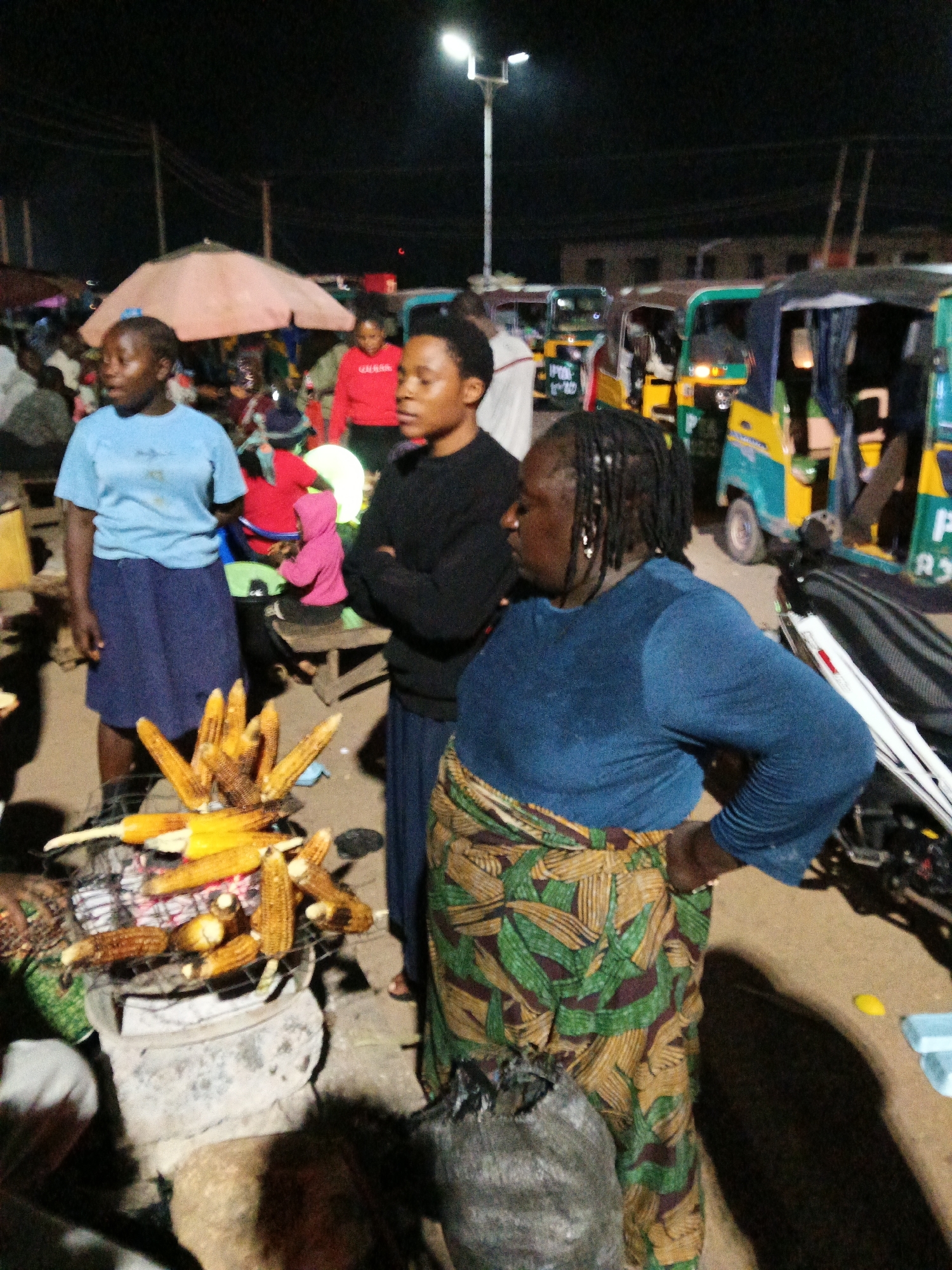 In Bukuru, Nigeria, at nighttime, a street food vendor is seen grilling corn. The vendor, a woman with dark skin and braided hair, wears a blue long-sleeved shirt and a patterned skirt. She stands next to a metal grill piled high with corn on the cob. Behind her, two young women are visible. One, wearing a light blue t-shirt and a dark blue skirt, stands to the left, while another, in a black long-sleeved shirt, stands to the right. The background reveals a bustling street scene with several brightly colored auto-rickshaws, commonly known as "keke napep," parked in a line. A large umbrella, possibly for shade or shelter, is also present in the scene. The ground appears to be dirt or unpaved road. The lighting suggests artificial illumination from streetlights and possibly the rickshaws themselves.