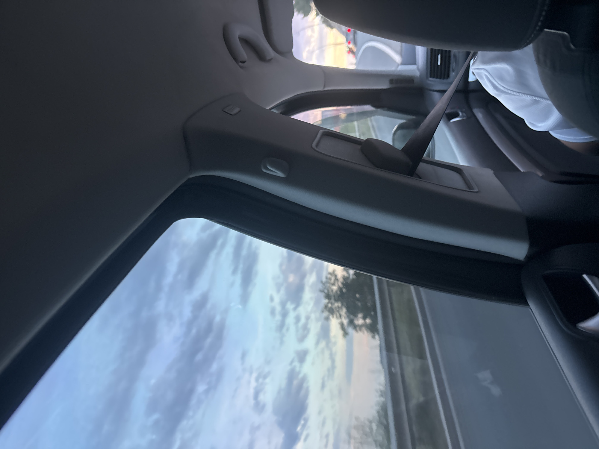 The image depicts a view from the interior of a moving vehicle, specifically from a rear-left passenger seat, looking out the left window and partially towards the front of the car, located in Kamenovo, Montenegro.
Outside the vehicle, a highway or major road is visible with a metal guardrail running alongside it. Beyond the road, green trees and vegetation line the landscape. The sky, a dominant feature, displays a mix of blue, grey, and faint pink or orange hues, suggesting a dusk or dawn setting with scattered cloud formations. Red taillights of other vehicles are visible in the distance, indicating traffic ahead on a curving road.
Inside the vehicle, the dark grey interior components are prominent. The B-pillar (door post) is visible with a seatbelt adjuster and an "AIRBAG" indicator. A dark seatbelt extends diagonally across the mid-right portion of the frame. The grey fabric roof liner is visible at the top, along with a grab handle. The back of a dark front passenger seat headrest is on the far right, partially obscuring a person wearing a white garment, likely a shirt or hoodie, in the front passenger seat. The lower part of the left door panel is also visible with a handle recess. The motion blur of the outside environment confirms the vehicle is in transit.