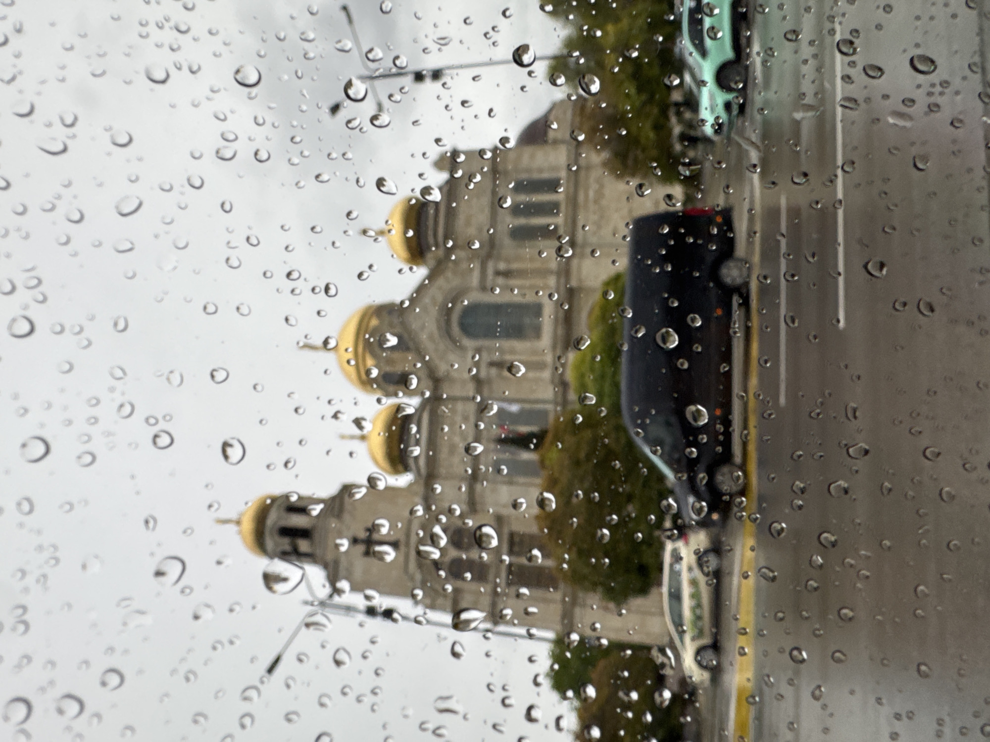 The image depicts a view through a rain-streaked window. Outside the window is the Dormition of the Mother of God Cathedral in Varna, Bulgaria. There are several cars parked on the street next to the cathedral. The sky is overcast. The image is oriented vertically.