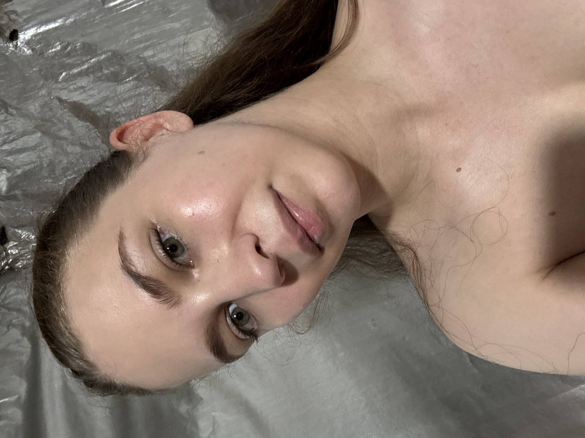The image is a close-up, head and shoulders selfie of a young woman with long brown hair pulled back. She has light skin with some redness on her cheeks and chest. Her eyes are green, and her lips are slightly parted and appear glossy. The background is a textured, reflective surface, possibly plastic sheeting. The lighting suggests an indoor environment, but the specific location in Nizhnyaya Gar', Russia, cannot be determined from the visual information provided. There are no other people or objects of note in the image.