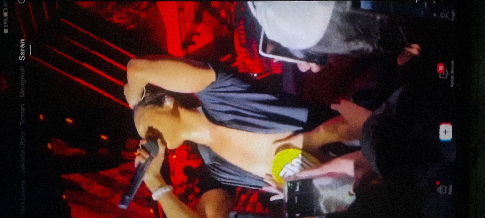 The image shows a close-up of a person singing into a microphone on a stage. The performer, a young male with light skin, is wearing a dark open-collared shirt that reveals his chest and abdomen. He has sweat on his skin and is holding a white beaded bracelet on his left wrist. His right arm is raised, and he is touching his head. In the foreground, a bright yellow object with some text on it is visible, held by hands in the audience. The background features red stage lighting and some blurry figures. The top of the image shows a navigation bar with text in Indonesian, including "Mini Drama," "Jakarta Utara," "Teman," "Mengikuti," and "Saran," which indicate that the scene is likely related to content on a social media platform and might be geographically tagged to North Jakarta, Indonesia. The overall environment appears to be a concert or live performance.