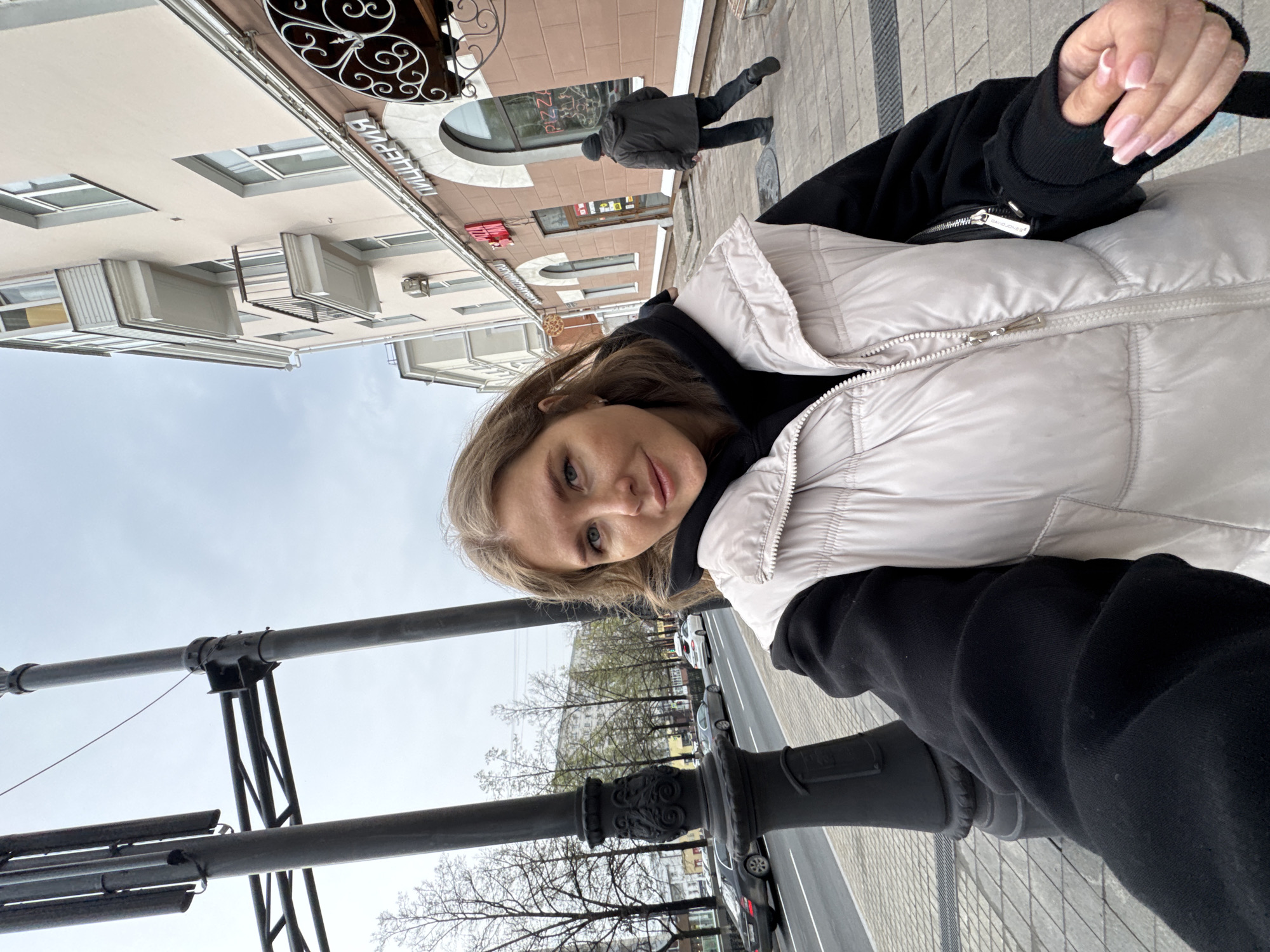 A woman in a white puffer vest over a black hoodie is taking a selfie in what appears to be a city setting. The location is possibly Perm, Russia, based on the architectural style of the buildings and the storefront signage in Cyrillic script. In the background, pedestrians are walking along a sidewalk, and cars are parked along the street. A black lamppost is in the foreground, partially obscuring the view of the street. The sky is overcast.