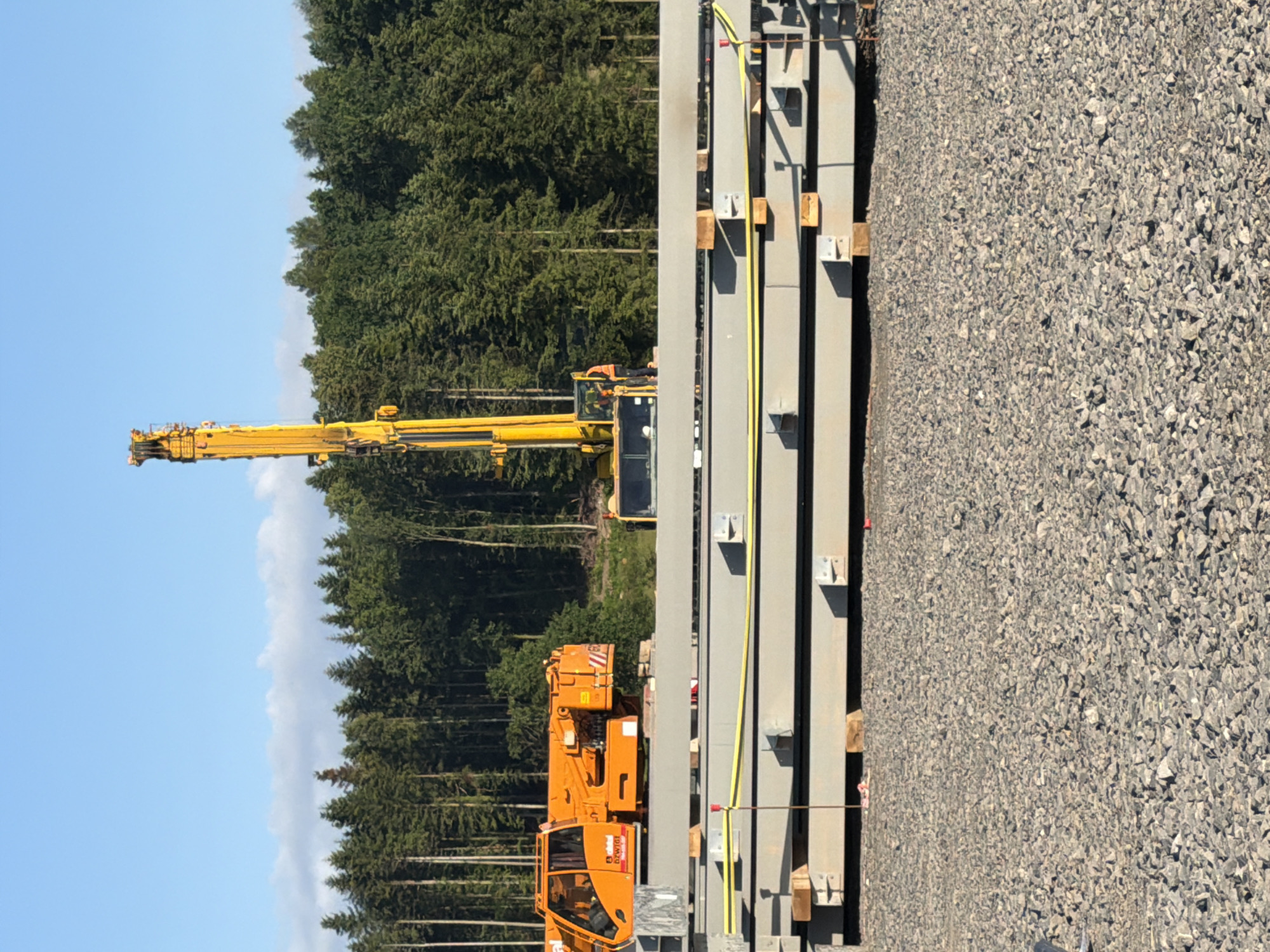 In Stróża, Poland, a construction scene unfolds under a clear blue sky with scattered white clouds. The foreground is dominated by a large pile of grey gravel. Stacked on top of the gravel are several large, rectangular concrete beams, each appearing to be several meters long. Yellow straps are visible, securing some of these beams.
In the middle ground, an orange mobile crane is positioned to the left, its cabin and boom clearly visible. To the right of the mobile crane, a yellow drilling rig stands tall, its long, slender mast extending upwards. A person in an orange vest and a helmet stands near the base of the drilling rig, appearing to be operating or supervising the machinery. Behind the construction equipment, a dense forest of tall green trees forms the background. The overall impression is one of ongoing construction work in a rural or semi-rural setting.