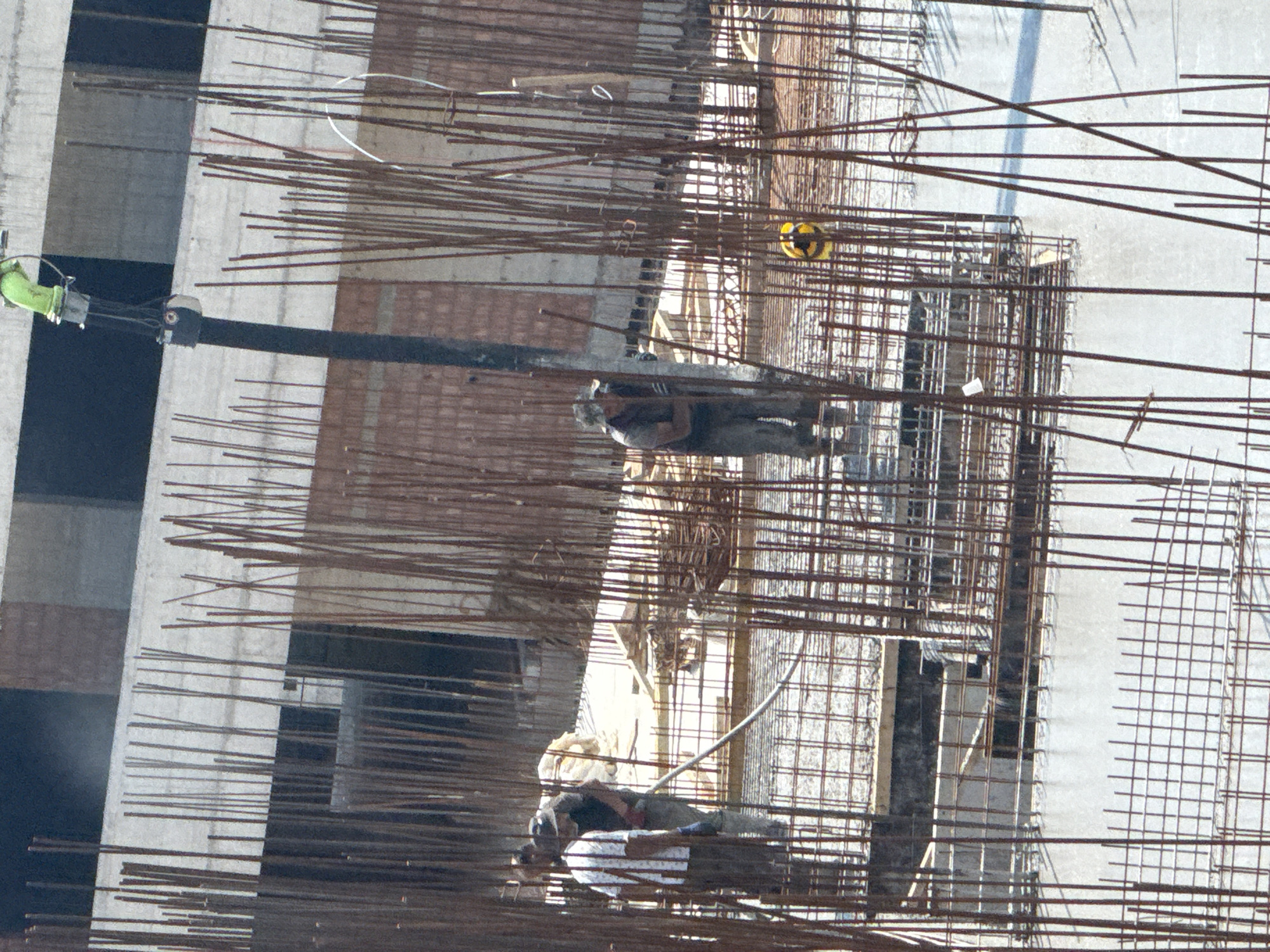 This media file shows a construction site in Boreti, Montenegro. The scene is dominated by a partially constructed building with exposed concrete walls and numerous metal rebar structures. Several people are visible on the site, appearing to be construction workers.
In the upper portion of the image, a worker wearing a yellow hard hat is partially visible. Further down, another worker in a dark shirt and pants is seen. Towards the bottom of the image, two more individuals are working, one wearing a white shirt. They appear to be engaged in construction-related tasks, though the specific activity is obscured by the rebar.
The lighting suggests it could be daytime, but the exact time is not discernible. The overall atmosphere is one of ongoing construction. There are no visible text elements in the image. The presence of rebar, concrete, and scaffolding clearly indicates a building construction project. The perspective is from above, looking down onto the site.