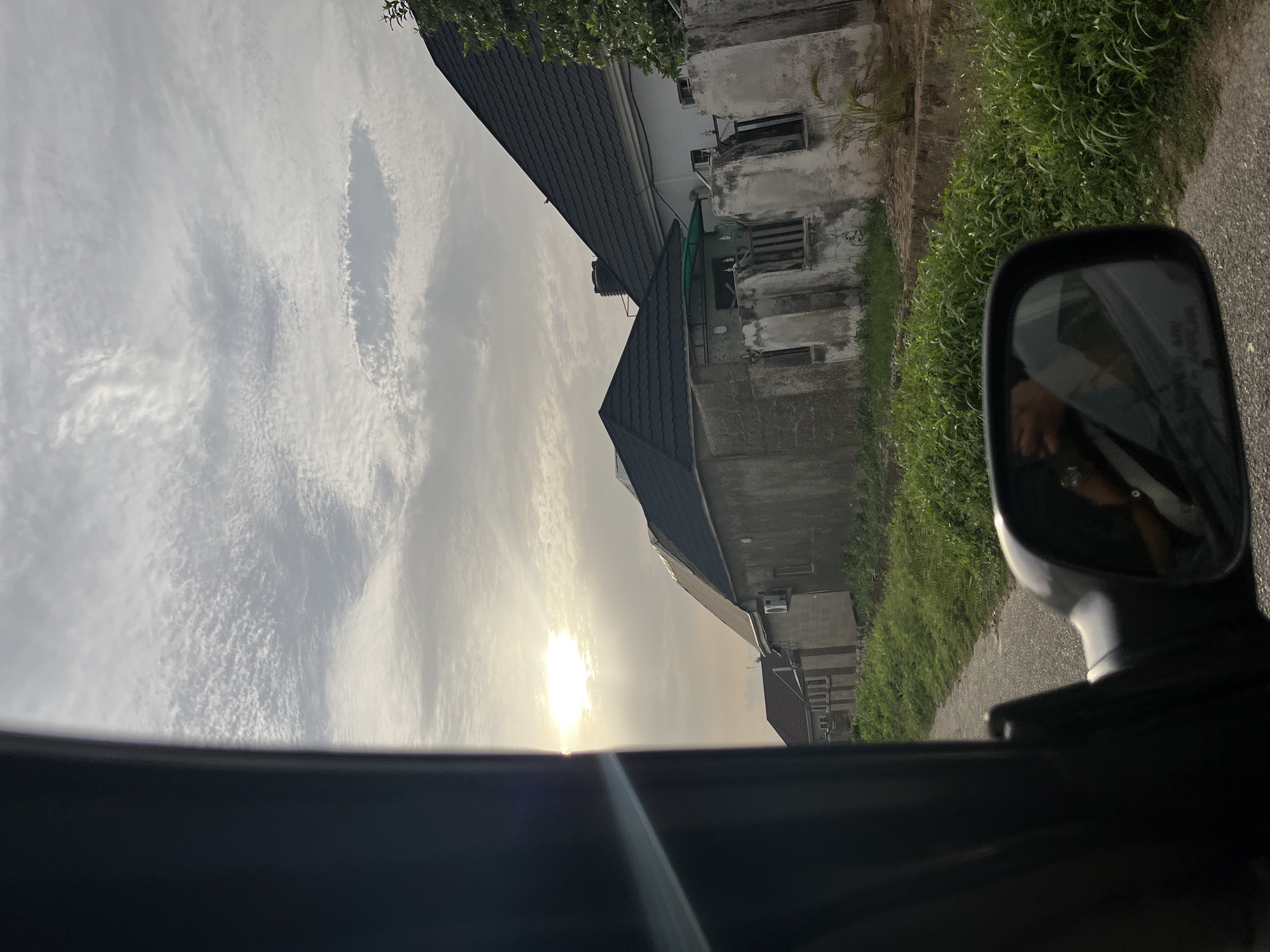 A sun-drenched scene viewed from inside a vehicle in what is possibly Abuja, Nigeria. The sun shines brightly through a partly cloudy sky. Several buildings of varying architectural styles, but sharing a similar weathered facade, are visible. Green vegetation borders what appears to be a road. A side mirror of the vehicle reflects a person inside. The car doorframe is prominent in the foreground.