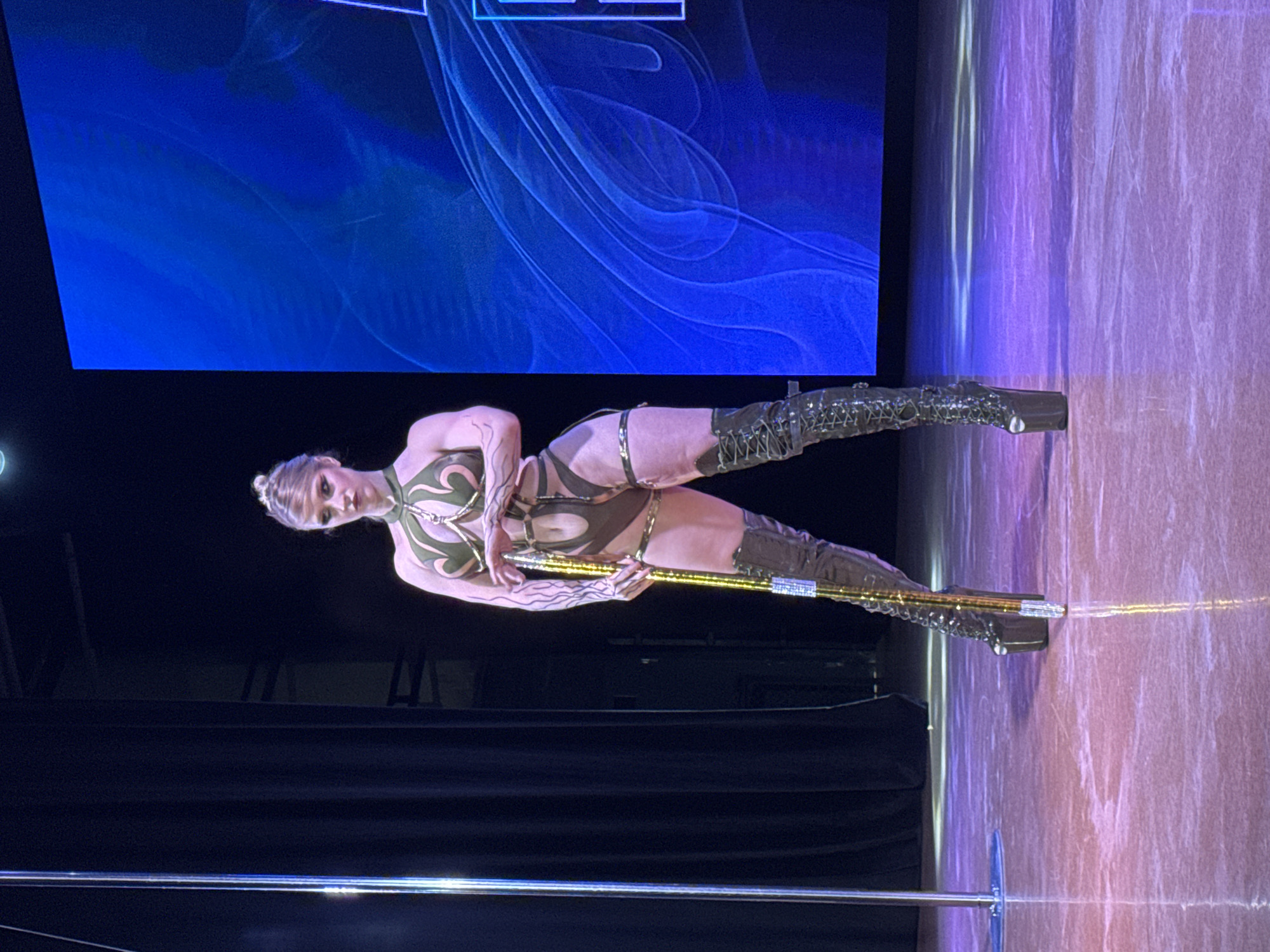 A blonde woman stands on a stage, holding a golden pole. She is wearing a dark, form-fitting costume with cutouts and thigh-high, lace-up boots. She has dark makeup on her eyes and her hair is pulled up. A blue digital screen is behind her, and a black curtain is to her left. There is a chrome pole to her left. The floor is a polished, reflective surface that is lit with purple and pink lights. This image was taken on July 14, 2024, in Krasnodar, Russia.