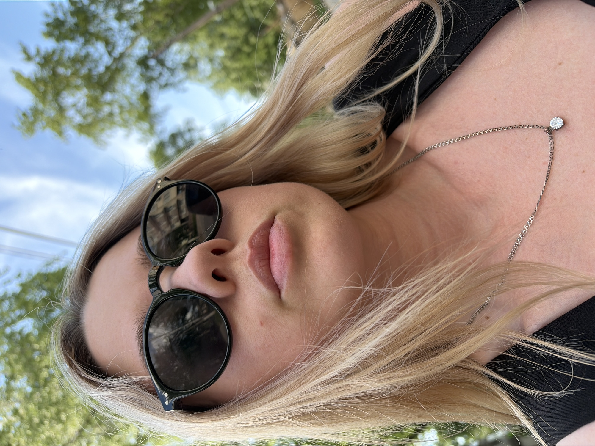A fair-skinned woman with blonde hair and sunglasses is the subject of this close-up, angled shot. Her lips are slightly parted. A silver chain necklace with a single diamond is visible on her chest. The background shows blurred green foliage, suggesting a park or outdoor setting. A fragment of the sky is also visible.
While the image may have been taken in Chaikovsky, Russia, there's nothing in the image to suggest this. The location can not be confirmed.