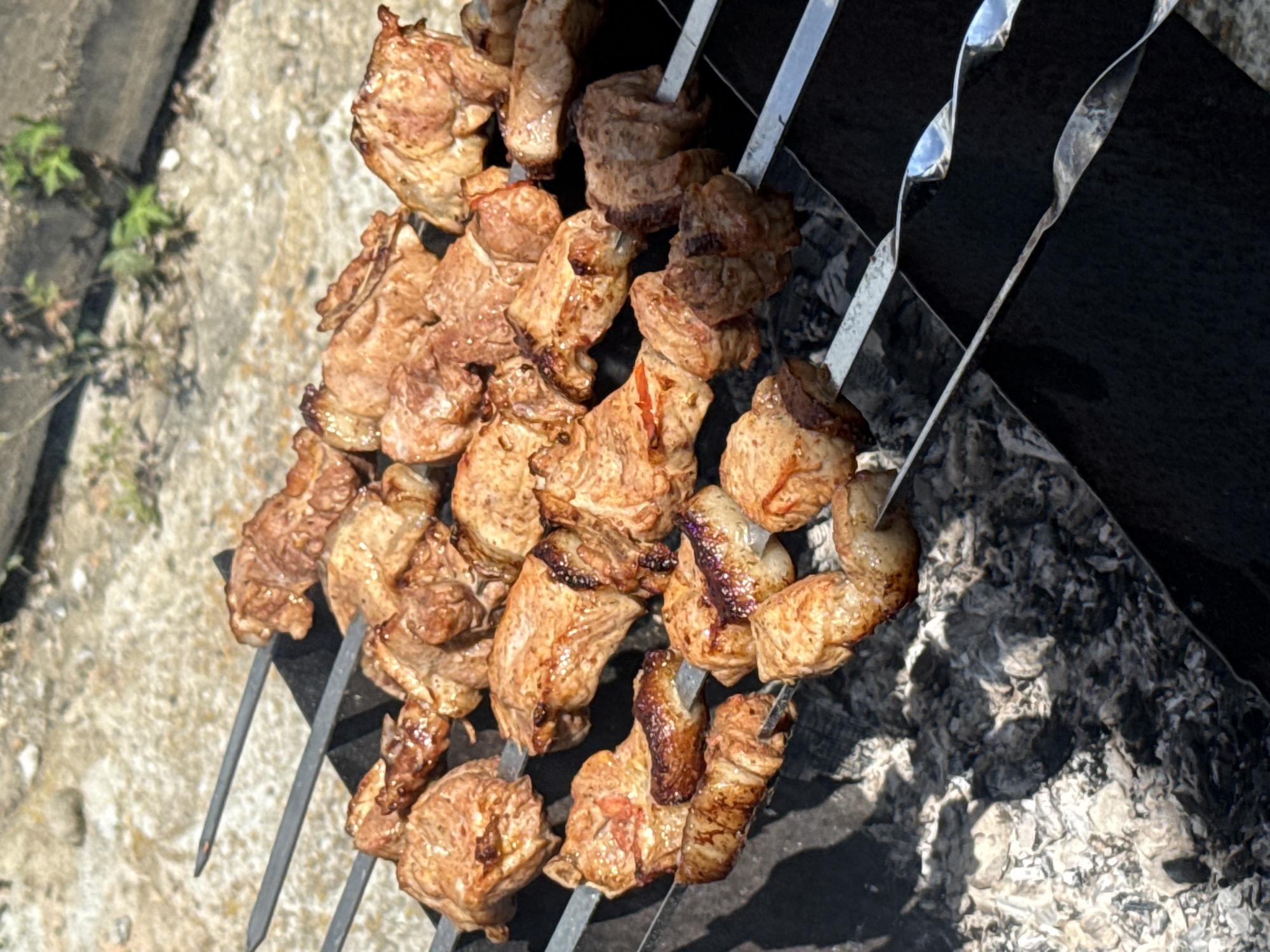 The image shows shashlik, a type of kebab, being grilled over hot coals. The meat is skewered and arranged on metal skewers, with several skewers visible in the foreground and middle ground. The shashlik appears to be well-cooked, with some pieces showing a browned and slightly charred exterior. The background is blurred but suggests an outdoor setting with gravel and vegetation. The scene is likely in Uyskoye, Russia, given the prompt. The focus is on the food being prepared, implying a culinary or recreational activity. There are no people or other animals visible in the image.