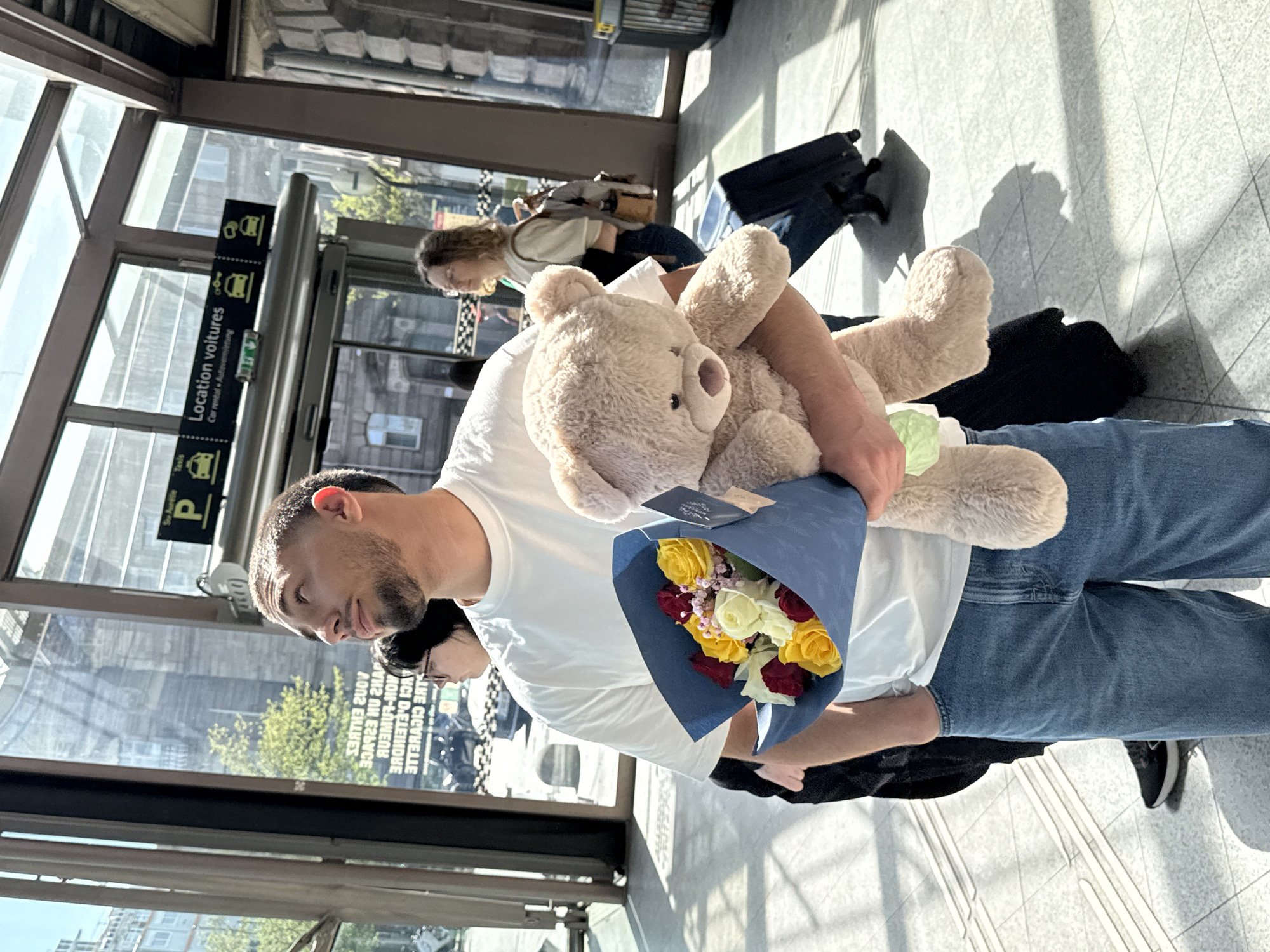 A man in a white t-shirt and jeans holds a bouquet of roses and a teddy bear inside the Gare de Strasbourg, a railway station in Strasbourg, France. He is standing near the "Location voitures" (car rental) sign. Other people and luggage are nearby, and sunlight streams through the glass windows.