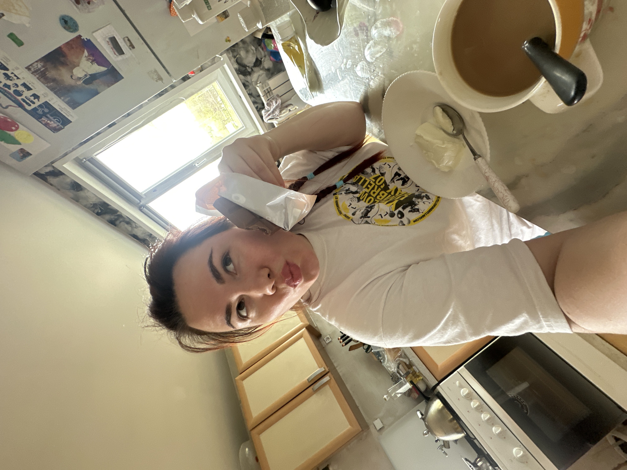 The image depicts a young woman in a kitchen. She is holding a small package, possibly of creamer, and posing for a selfie. On the counter are a mug of coffee with a spoon, and a plate with a dollop of what appears to be cream or butter. The kitchen has light-colored cabinets and an oven. A window provides natural light. There is no verifiable information to determine the location is Черёмушки, Russia.