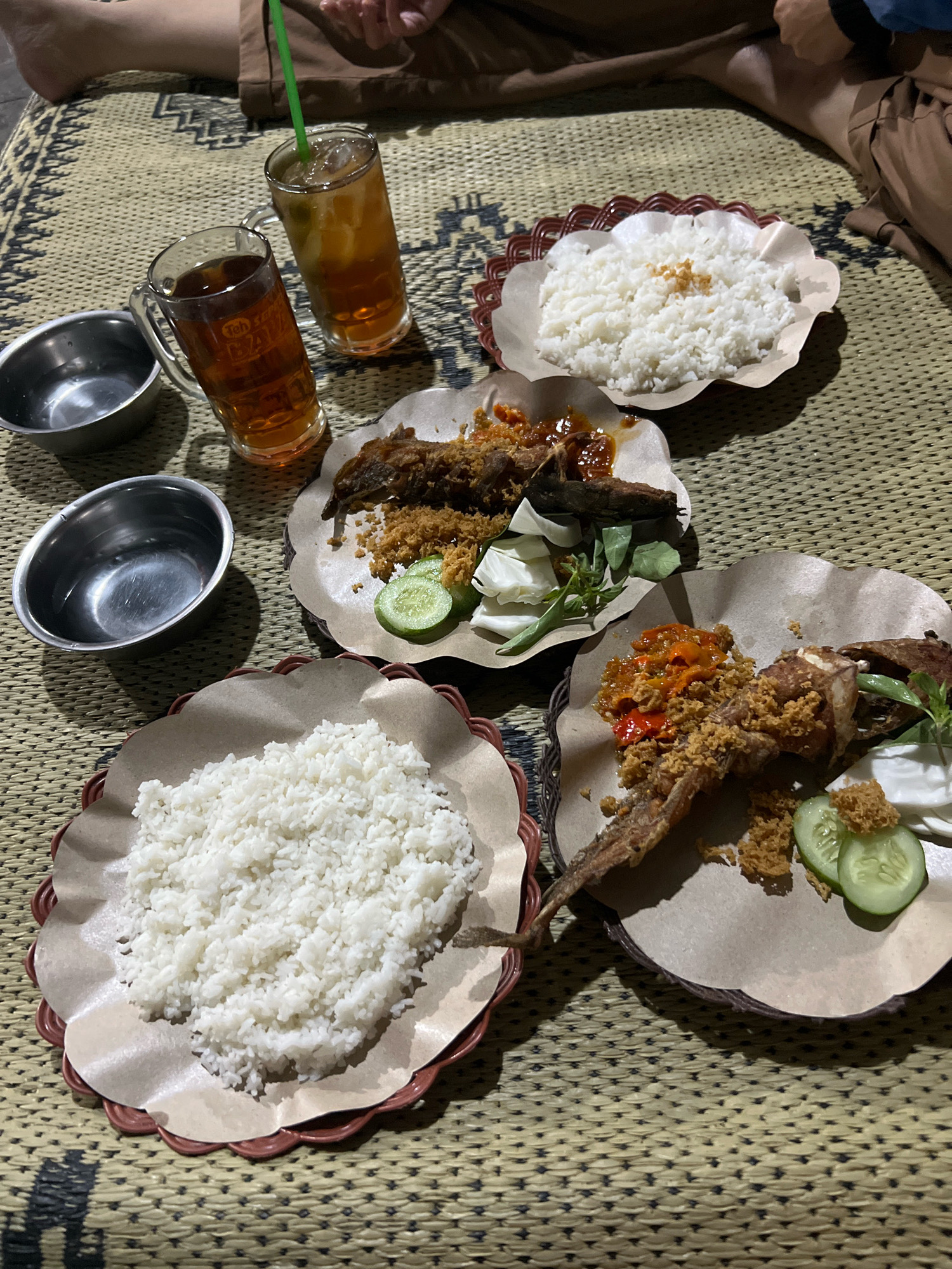 The image shows a dimly lit outdoor setting with several plates of food and drinks arranged on a woven mat. Two glasses of iced tea are visible, one with a green straw. There are two metal bowls and three plates of food. Each plate contains rice, fried chicken or fish, and some vegetables, possibly cucumber and cabbage. One plate has a serving of sambal or spicy sauce. The food appears to be served on paper liners within decorative, red-rimmed plates. The background shows parts of people's legs, suggesting they are seated on the mat. The overall scene suggests a casual outdoor dining experience. The location is Tlogoadi, Indonesia, a village known for its rice fields and traditional Indonesian cuisine.