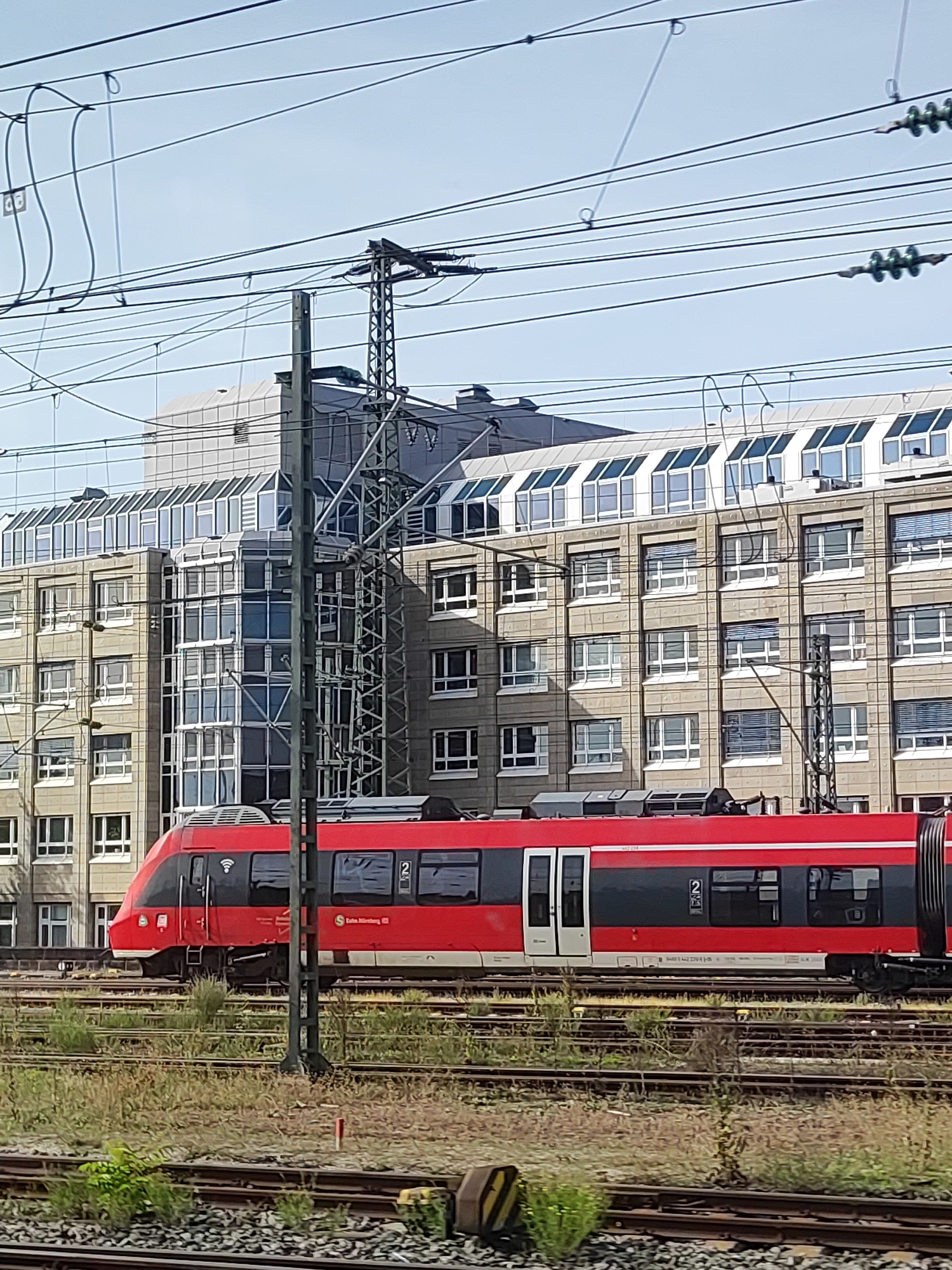 A red and black passenger train is seen from the side, moving on railway tracks in front of a modern office building. The building has multiple floors with rows of windows and a distinctive glass roof structure. Overhead, a complex network of overhead electrical wires for the train is visible, supported by metal poles. The area around the tracks is overgrown with grass and weeds. The scene appears to be an urban setting, likely near a train station or railway yard. The train has the number "2" displayed on its side. The building features a stone facade.