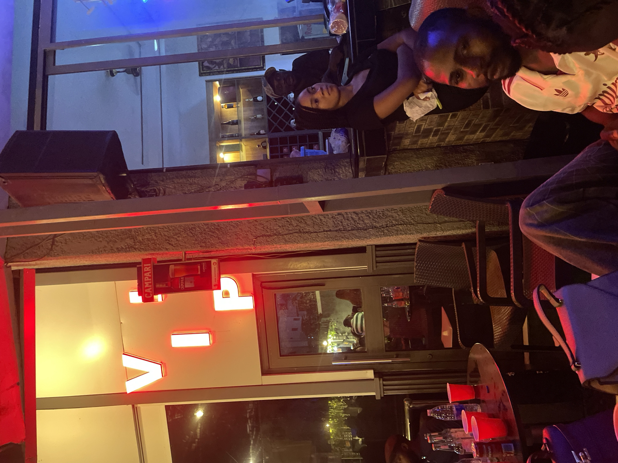 The image depicts a nighttime scene at a bar or club, likely in Guzape, Nigeria, as indicated by the "VIP" sign. The foreground features a round black table with several red plastic cups, bottles, and possibly a cooler. Two purple handbags are placed on the floor in the lower left and center. A man in a white shirt with red stripes is seated in the lower right, looking towards the camera. To his left, a woman in a black shirt is seated, facing away from the camera. Behind them, a man in a dark shirt and a cap stands at a bar area. The bar has shelves stocked with bottles and a wine rack. In the background, through a glass door, several people are seated at tables, and the exterior of a building with yellow lighting is visible. A large speaker is mounted on a pillar to the left of the bar area. Neon red lights illuminate the "VIP" sign, which is mounted on a white wall above the entrance. The overall atmosphere appears to be casual and social, with ambient lighting contributing to a nighttime entertainment venue.