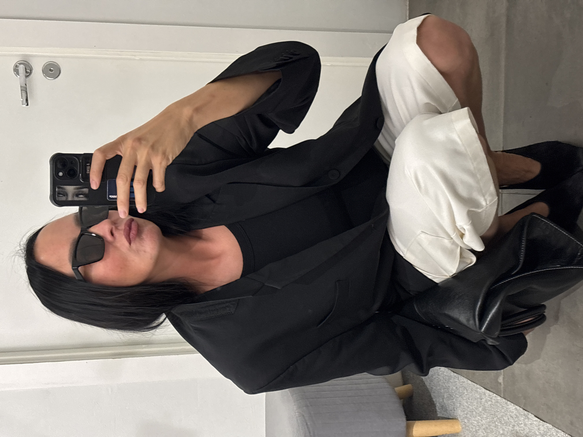 A person is taking a mirror selfie in what appears to be a fitting room or dressing area. They are wearing a black blazer over a black top, white shorts, and black shoes. They are also wearing black sunglasses and holding a black smartphone in their right hand. The background shows a white door with a silver handle and a gray upholstered bench. The floor is gray. The lighting is bright. There are no specific details in the image that would indicate the location being the city of Chaikovsky, Russia.