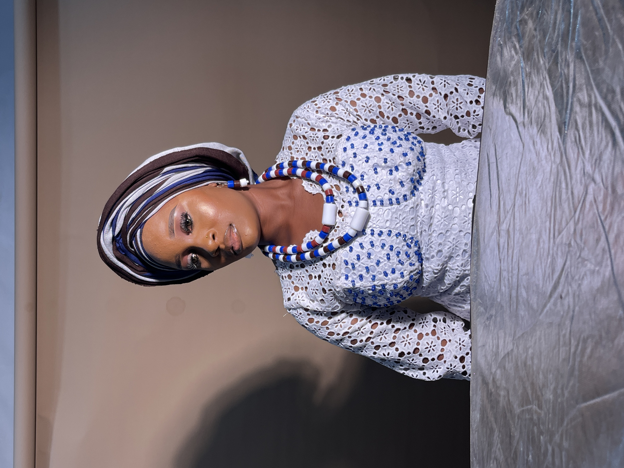 A woman is depicted in a medium shot, seated at a table covered with a shimmering silver cloth. She is wearing a white lace top adorned with blue beads and a multi-colored headscarf. She is also wearing a beaded necklace in red, white, and blue. The background is a plain, neutral color. The lighting suggests a studio setting with a shadow cast on the wall behind her. The image appears to have been taken indoors.