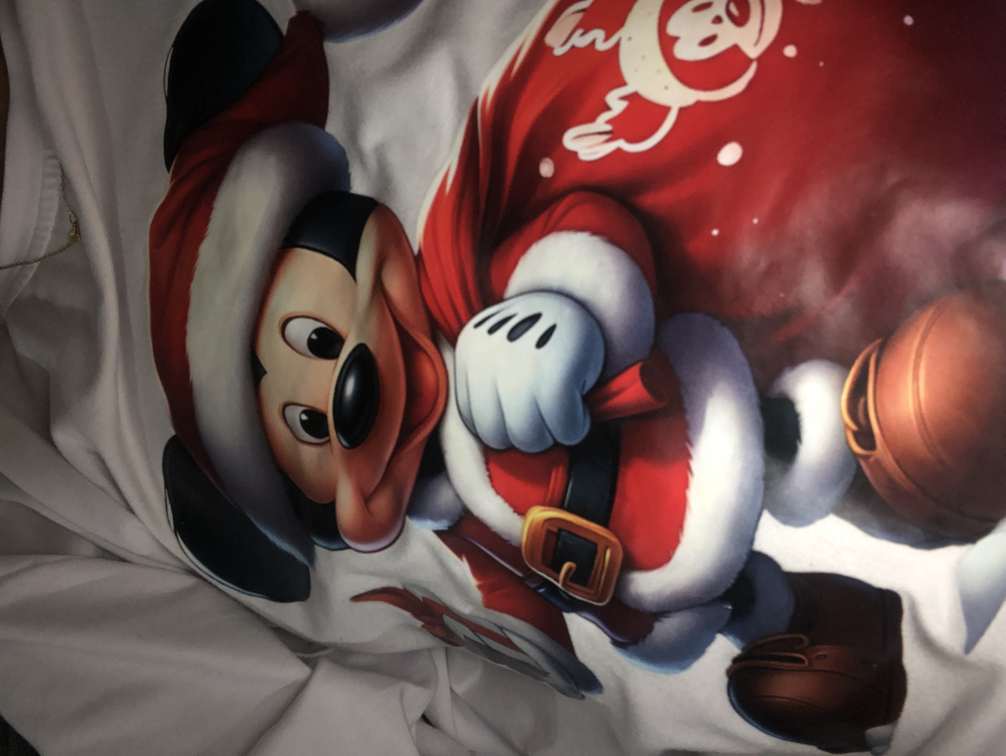 The image shows a close-up, high-angle, static shot of a white fabric with a detailed print of a cartoon character, Mickey Mouse, dressed in a Santa Claus costume. Mickey is depicted from the waist up, wearing a red Santa hat with white trim, a red coat with white fur trim, white gloves, and brown shoes. He is holding a red sack decorated with smaller images of Mickey Mouse wearing Santa hats. The fabric appears to be a t-shirt or similar garment. The background is a softly lit, white, and creased material, likely part of the same garment. There is no discernible connection to the city of l'Hospitalet de Llobregat, Spain.