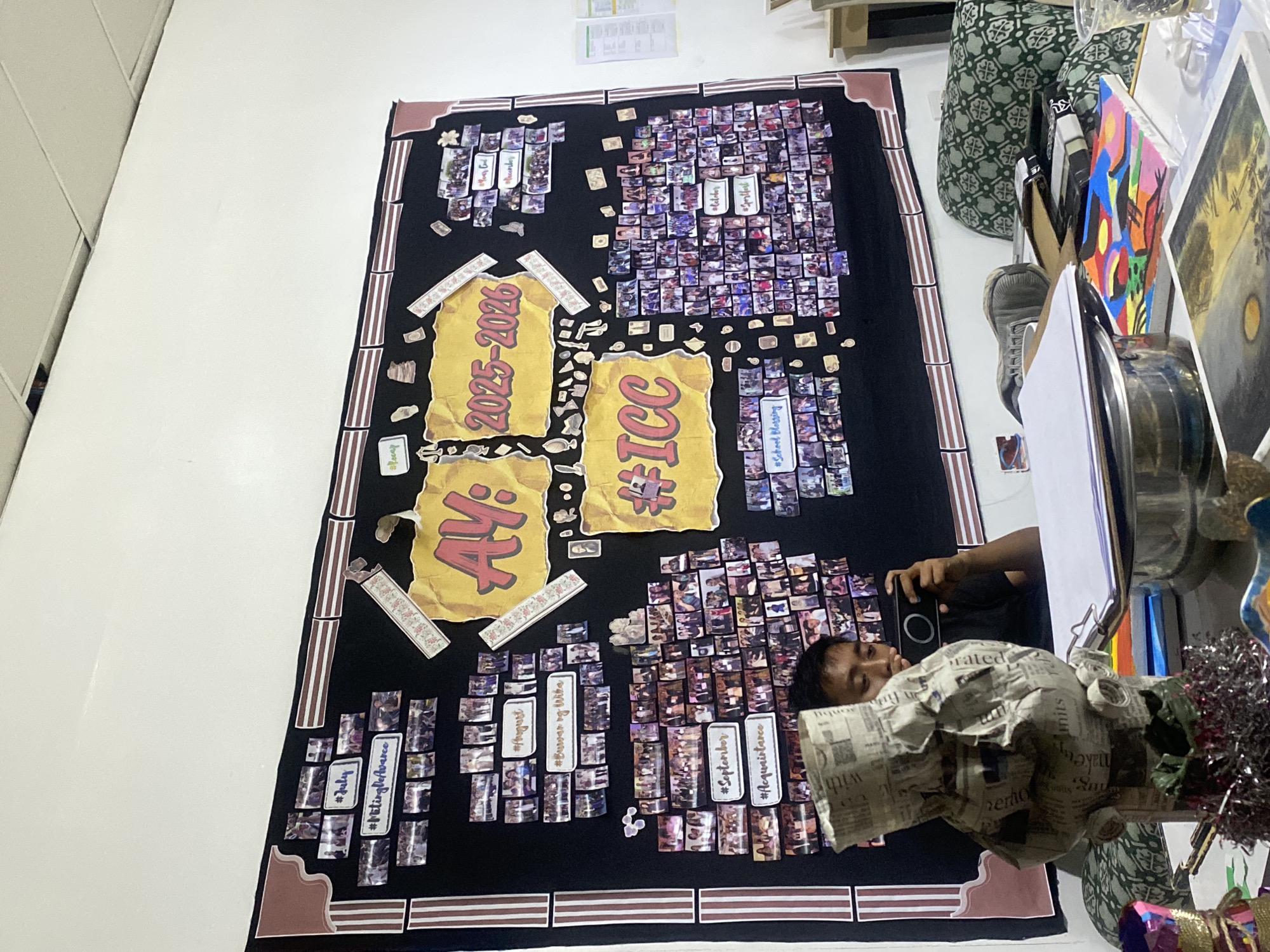 This image captures an indoor scene, primarily featuring a large, black display board adorned with numerous small photographs and text elements. The board is oriented vertically and appears to be mounted on a white wall, possibly in a school or event space. It has a distinctive brown and white striped border, reminiscent of a framed poster.
Central to the display are large yellow torn paper cutouts. One prominently displays "AY: 2025-2026" in bold red text, indicating an academic year. Another cutout reads "#ICC". The board is densely covered with small, square, vertically aligned photographs, arranged in clusters. These photos appear to be candid shots of individuals and groups participating in various activities, suggesting school or college-related events. Interspersed among the photo clusters are rectangular labels with hashtags and month names, categorizing the images. Visible text on these labels includes "#July", "#MitingDeAvance", "#August", "#BuwanNgWika", "#September", "#Acquaintance", "#October", and "#SchoolBlessing".
On the right side of the image, a man is visible, looking intently at the display and holding a black digital camera, seemingly taking a picture. Partially visible beside him are various objects, including a green patterned cushion, several framed artworks (one abstract and colorful, another depicting a landscape with a large yellow sun), and a reflective silver object. In the lower right foreground, a sculptural object wrapped in newspaper, with visible text snippets like "hermo" and "make," adds to the cluttered, creative environment. The overall impression is a vibrant commemorative display, likely celebrating a recent academic year's events at an institution in Davao City, Philippines.