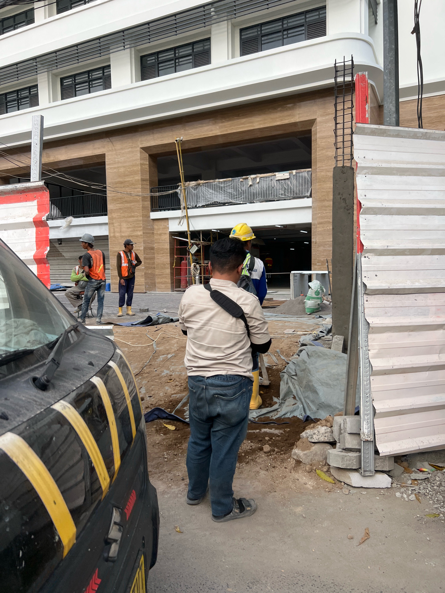 The image shows a construction site in the city of Yogyakarta, Indonesia. In the foreground, a black vehicle with yellow stripes is parked on the left. Three construction workers are visible in the middle ground, two of whom are wearing safety vests and hard hats. One worker is standing while the other two are seated or partially visible. In the background, a building under construction is partially visible, with scaffolding and construction materials present. The ground is covered with dirt and debris. The overall scene depicts an active construction area.