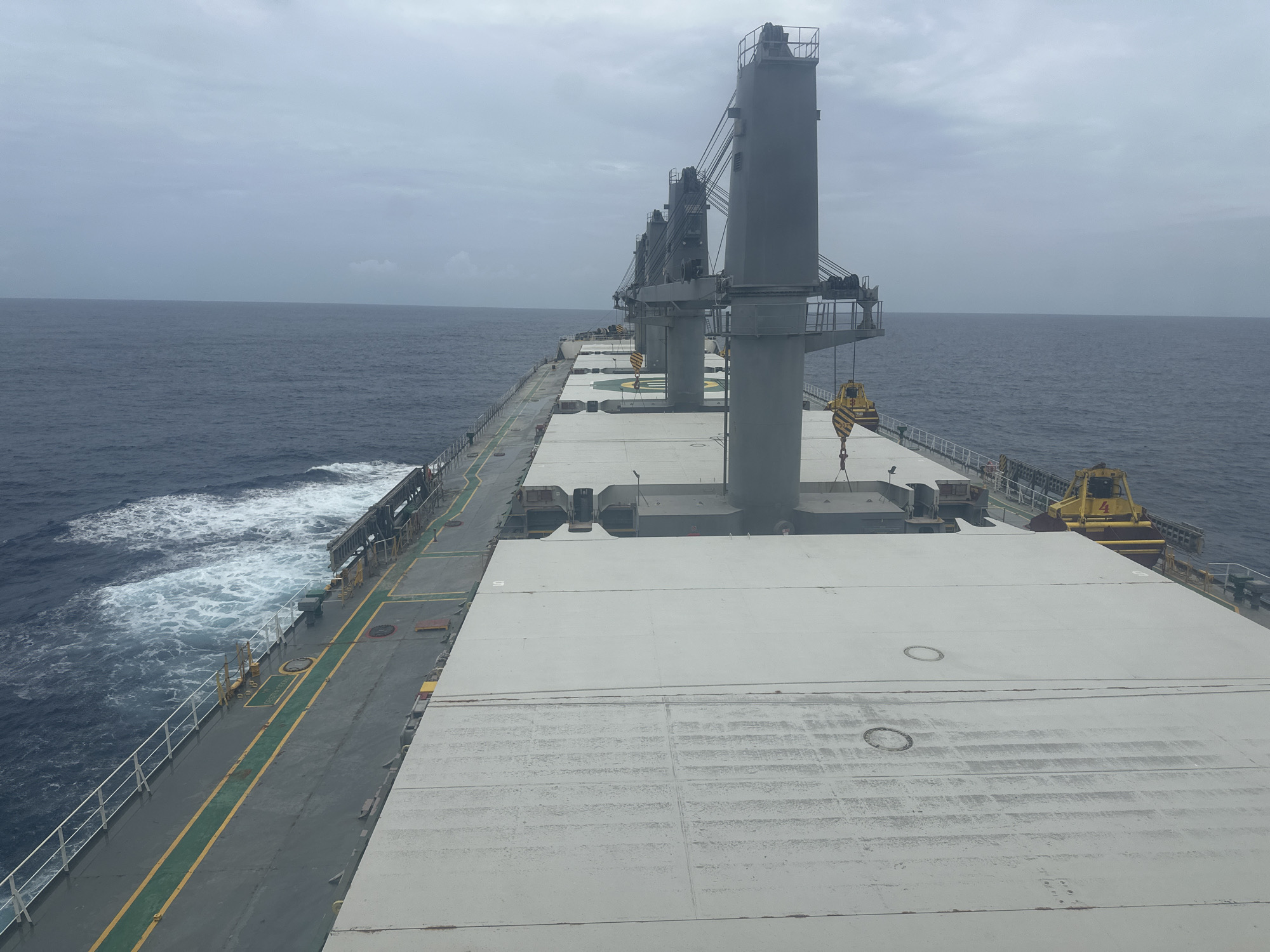 This media file depicts a perspective from the deck of a large cargo ship at sea. The dominant subjects are the ship itself, with its prominent grey cranes and the expanse of its white, panelled deck, and the vast, dark blue ocean stretching to the horizon under an overcast sky. The setting is clearly maritime, with the ship moving through the water, evidenced by the foamy wake trailing behind its port side.
The ship's deck is mostly empty, save for the massive, tall cranes that line its length, suggesting it is equipped for loading and unloading cargo. The cranes are grey and metallic, with complex rigging and pulleys. One yellow hook is visible, hanging from a crane. The deck itself is a light grey, with some visible wear and tear, including streaks and circular markings. Green and yellow lines mark walkways and safety zones along the port side. There are also circular hatches visible on the deck.
The weather appears overcast and possibly gloomy, with a uniform grey sky and a muted light. The sea is calm but active, with waves creating white foam. No people are visible in the frame, reinforcing the sense of the ship being at sea, perhaps on a long journey. The overall mood is one of quiet travel and industrial scale.
There is no visible text in the image, except for a "4" painted in white on a yellow piece of equipment on the starboard side, near the stern of the ship. The time of day is indeterminate due to the overcast conditions, but it appears to be daytime. The location is unknown, but the open ocean is clearly the environment.