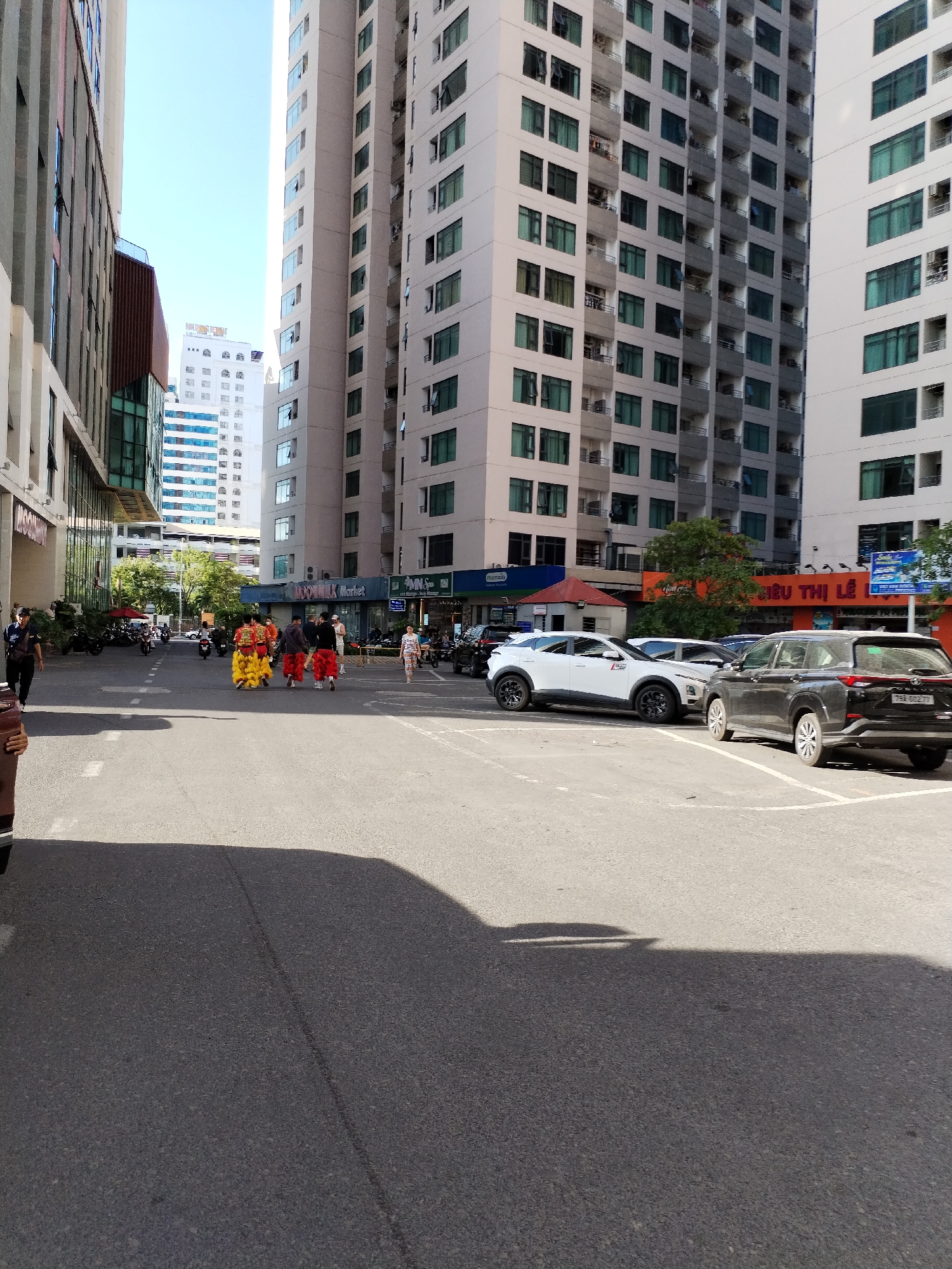 The image depicts a street scene in Bac Nha Trang Ward, Vietnam. Several tall buildings dominate the background. In the foreground, there is a street with parked cars. A group of people dressed in traditional lion dance costumes are walking down the street. A black SUV and a white SUV are parked in the parking spaces. Pedestrians can be seen near the buildings and market. The lighting suggests it is daytime. The overall atmosphere indicates an urban setting with a cultural event taking place.