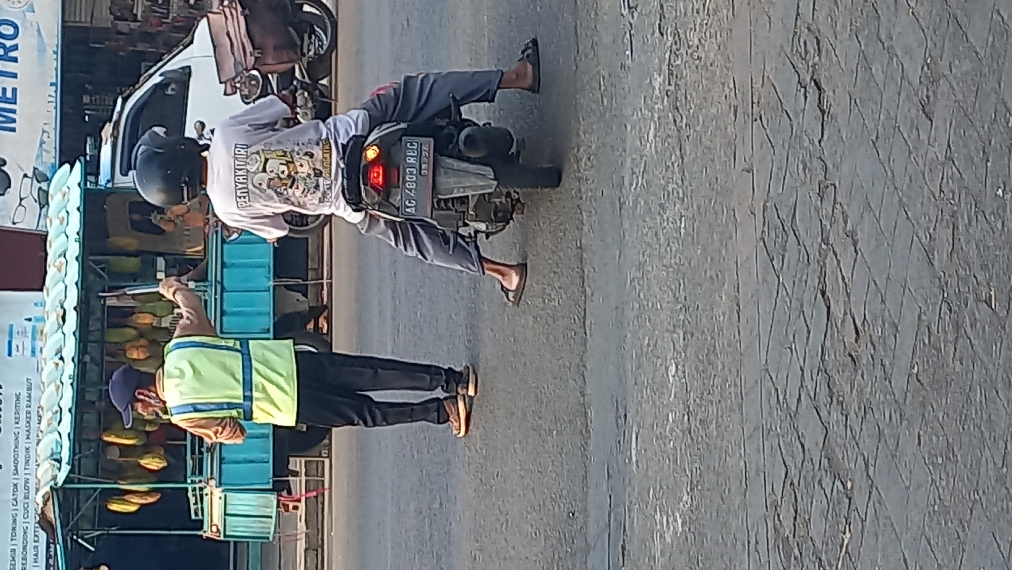 The image depicts a street scene in Mangunsari, Indonesia. In the foreground, a person wearing a bright yellow vest and a blue cap stands on the asphalt, directing traffic with an outstretched arm. They are wearing dark pants and brown shoes. To the right, another person is seated on a motorcycle, wearing a helmet and a light-colored t-shirt with a graphic print. The motorcycle has a license plate with the inscription "AC 4803 RBC". Behind the pedestrian, a mobile food stall with a green roof is visible, displaying a row of what appear to be durian fruits. A white vehicle is parked in the background, and the street is paved with asphalt. The overall environment suggests an outdoor urban setting, likely a market area or a busy street.