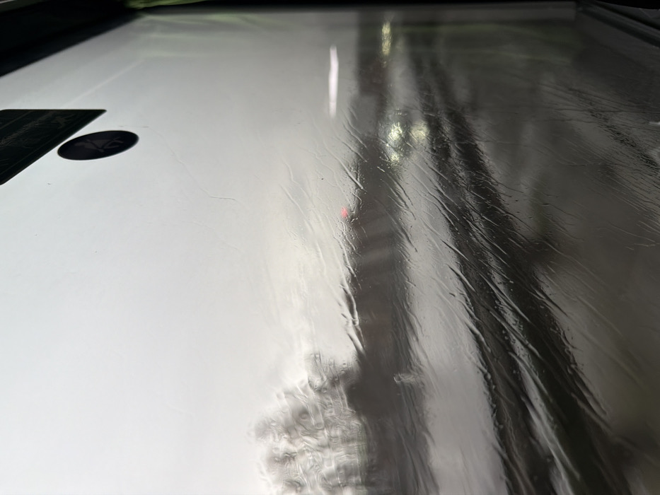 The image depicts an exterior scene viewed through a rain-streaked window, likely from a moving vehicle, in Moineville, France. The foreground is dominated by the window surface, covered in numerous vertical and diagonal rivulets of water, blurring and distorting the distant view. A dark rectangular object, possibly a sticker or label, is partially visible in the upper right corner, along with a smaller circular black sticker below it.
Beyond the window, the environment appears overcast and dim. A multi-lane roadway, slick with rain, occupies the midground and lower portion of the frame. Traffic is present, with at least one vehicle displaying illuminated white headlights on the right side of the road, and another exhibiting a red taillight or brake light in the center-left. Trees or dense vegetation, their branches appearing bare, are visible on the left side of the roadway, heavily blurred by the rain and atmospheric conditions. The sky above is uniformly grey and overcast. No specific individuals are discernible.