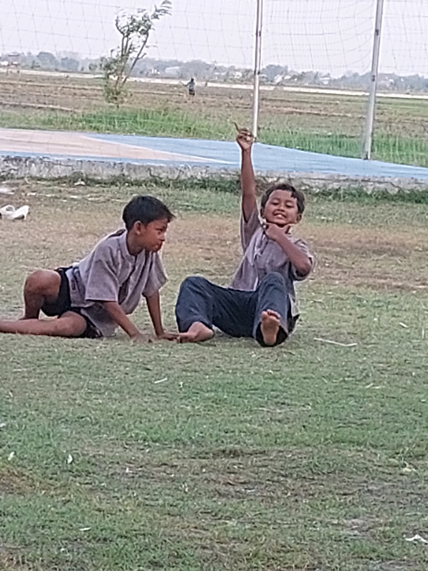 Two young boys are sitting on a grassy field near a track with a blue and white border. The boy on the left is wearing a grey t-shirt and dark shorts. He is on his hands and knees, looking to the right. The boy on the right is wearing a grey t-shirt and dark pants. He is sitting with his legs extended, holding one finger up in the air and smiling. In the background, there is a net, a tree, and a field with some cultivation. The environment appears to be outdoors, possibly a sports field or park, in Sarangan, Indonesia.