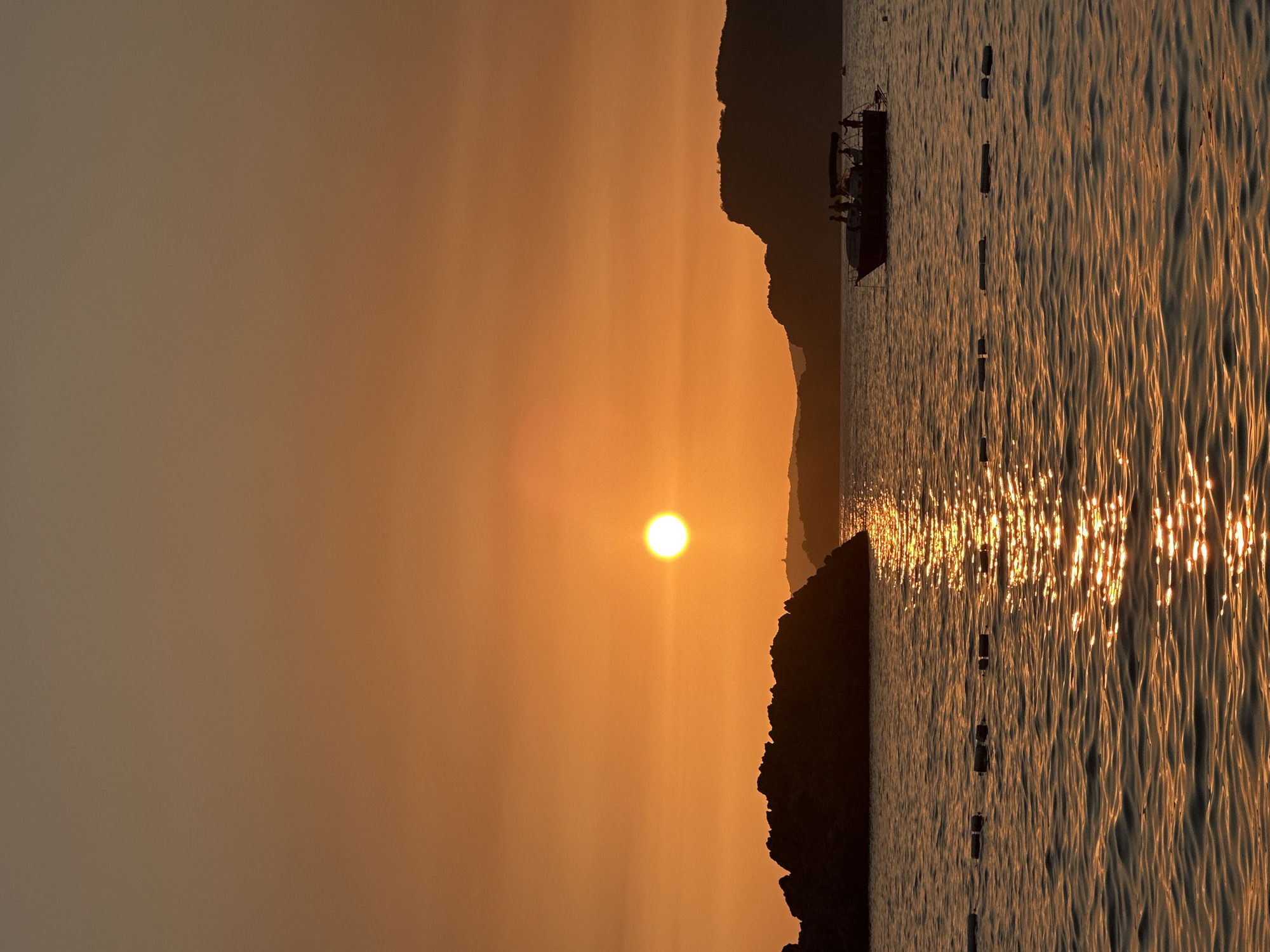 An outdoor, vertical, medium shot captures a sunset over the sea at Sveti Stefan, Montenegro. The sun, a bright white orb, hangs low in the hazy orange sky, casting a shimmering golden path across the rippling water. In the foreground, a line of buoys marks a swimming area. To the left, a large, dark rock formation juts out of the water. In the mid-ground, a boat with several silhouetted figures on board cruises on the sea. The background features the silhouettes of distant hills and mountains.