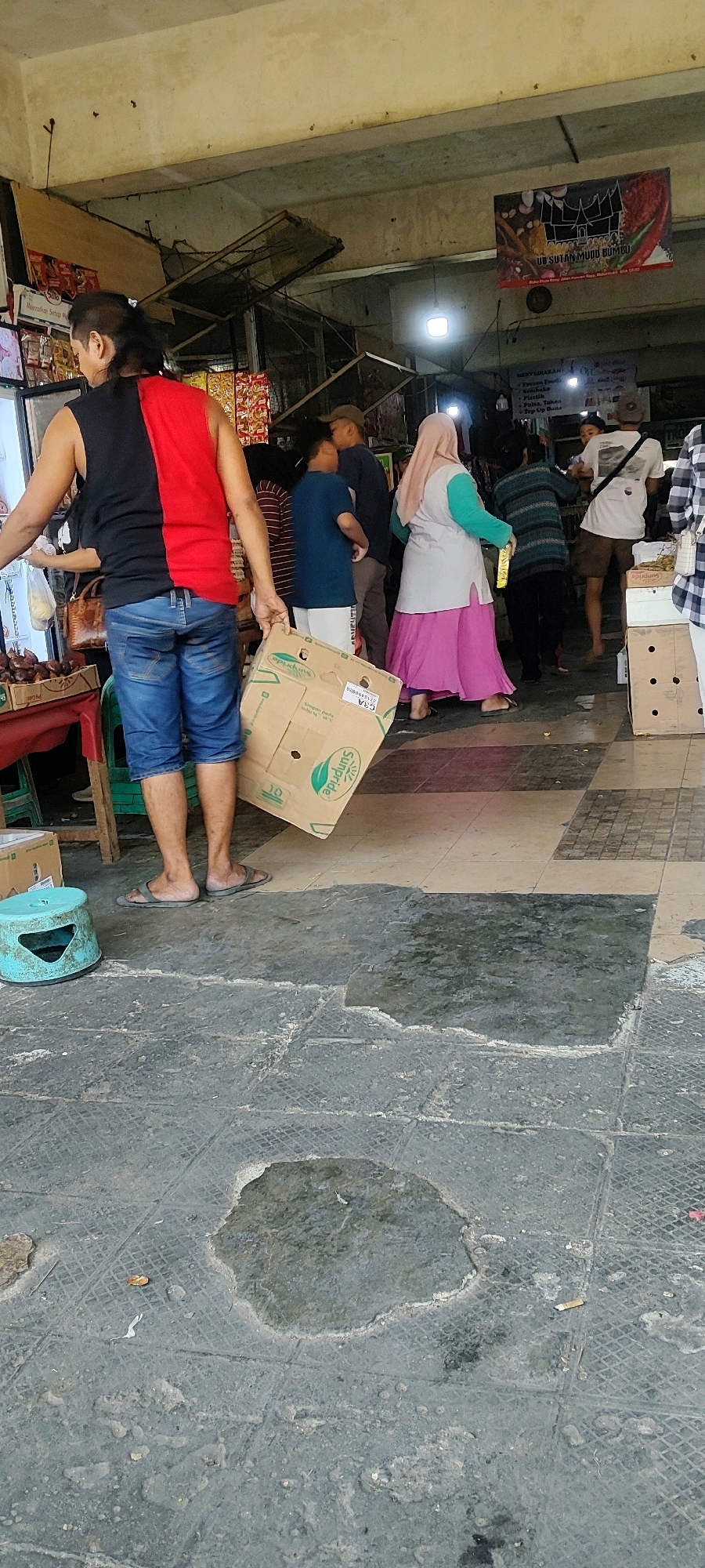 In an outdoor market setting in Mekarmukti, Indonesia, a man with dark hair pulled back in a ponytail is seen from the waist down, wearing a black and red sleeveless shirt and blue denim shorts. He is holding a flattened cardboard box with green lettering from "Sunpride". To his left, a teal-colored plastic stool is visible. The ground is paved with dark tiles, some of which are wet, creating a puddle in the foreground. In the background, several other people are milling about, some appearing to be vendors or shoppers. One woman wears a pink hijab and a long pink skirt, and another person in the background is wearing a striped long-sleeved shirt. The market appears to be covered by a roof, and there are signs and stalls visible.