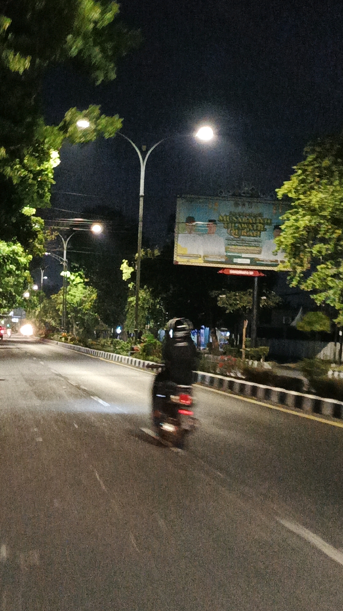 At night, a person on a scooter travels down a paved road in Surakarta, Indonesia. The scooter rider is wearing a dark helmet and jacket. The road is lined with trees and streetlights. A billboard is visible in the background, and the sky is dark.