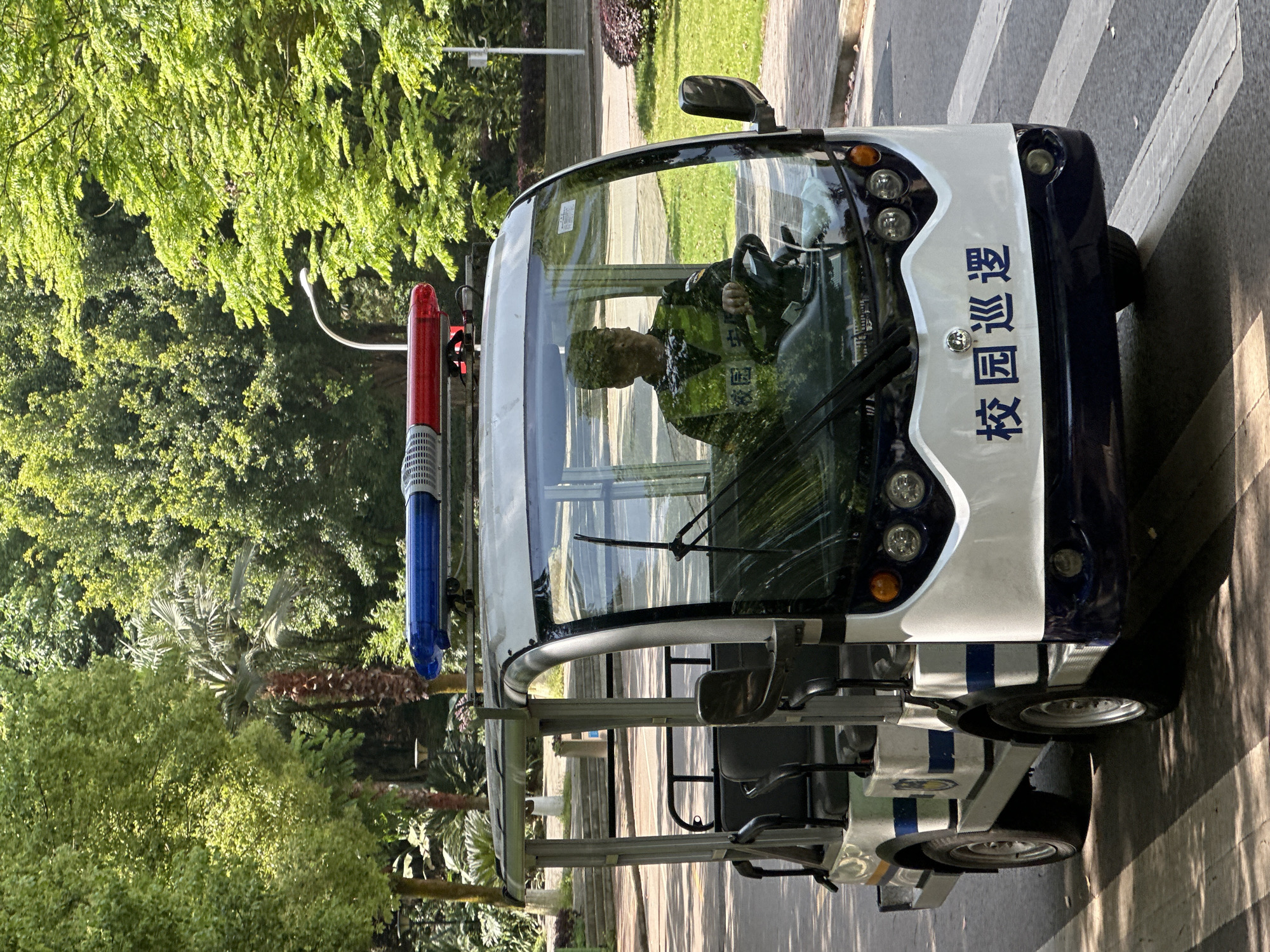 An outdoor scene in Jiangjin, China, shows a white and dark blue utility vehicle, likely an electric patrol cart, parked on a paved surface next to a pedestrian crosswalk. Lush green trees and foliage flank both sides of the pathway, with sunlight casting distinct shadows, indicating a bright, clear day.
The vehicle is the central focus, featuring a red and blue flashing light bar mounted on its roof. A person wearing what appears to be a camouflage-patterned shirt and a reflective vest is seated behind the steering wheel, visible through the windshield. The driver's reflection and the angle obscure their facial features. The vehicle has multiple headlights and a side mirror. The rear section of the vehicle appears to have additional seating, suggesting a capacity for more passengers.
Prominently displayed on the side of the vehicle, in dark blue Chinese characters on a white background, is the text "校园巡逻" (Xiàoyuán Xúnluó), which translates to "Campus Patrol." This identifies the vehicle's function and suggests the setting is a school or university campus. The vehicle itself has a clean design with horizontal blue stripes.
The activity captured is the vehicle being stationary, with its driver ready or paused in operation. The vibrant greenery and clear lighting contribute to a pleasant daytime atmosphere.