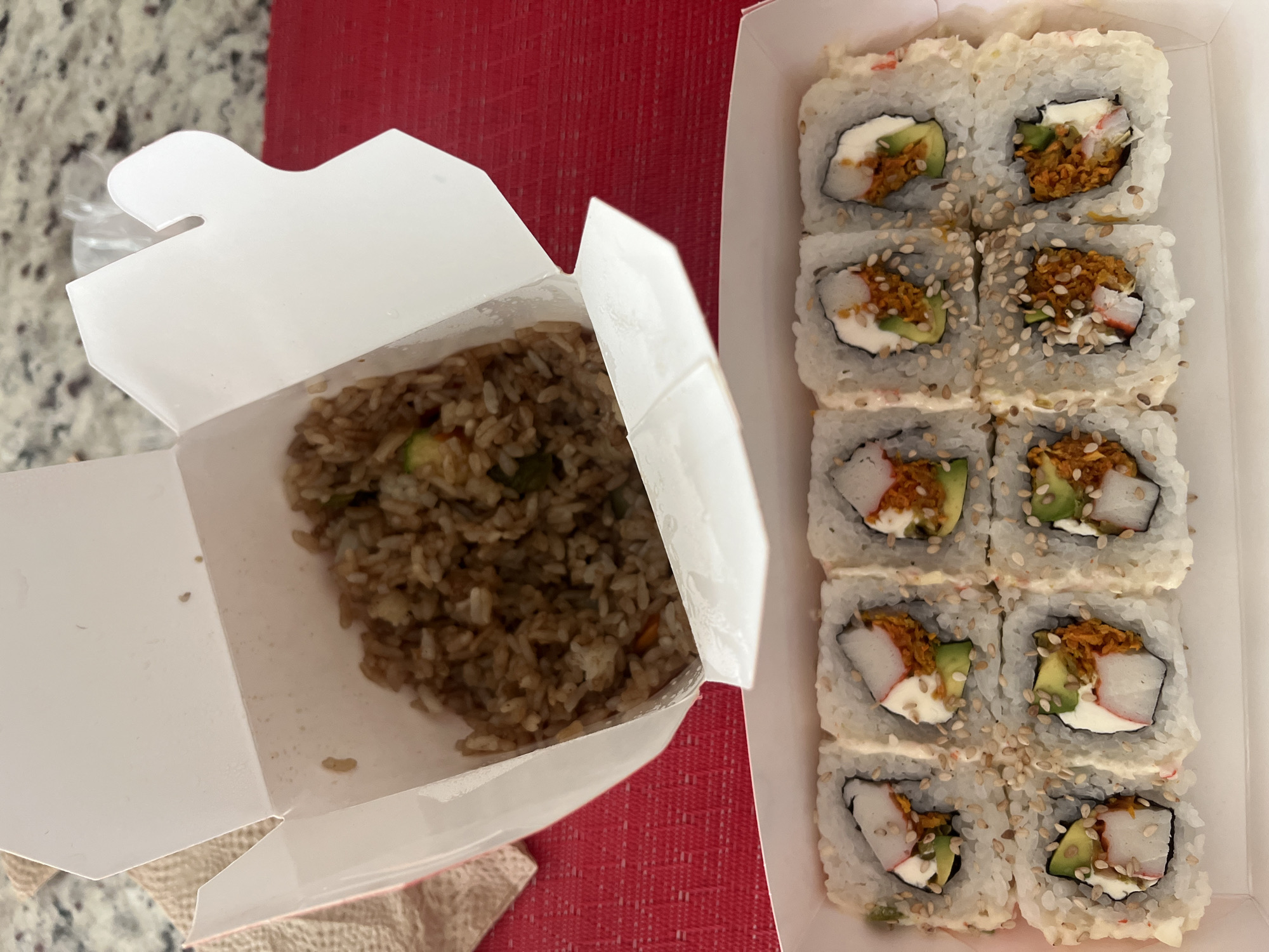 An overhead shot displays two open takeout containers filled with food, resting on a textured red surface, likely a placemat or tablecloth. A lighter, mottled countertop is visible in the top left background.
On the left, a partially open white Chinese takeout box contains a portion of fried rice. The rice is light brown with visible green pieces, likely vegetables, mixed in. The container's inside walls show some remnants of food.
To the right, a rectangular white cardboard container holds ten pieces of sushi arranged in two rows, with space for two more pieces at the bottom, suggesting an original total of twelve pieces. The sushi rolls are covered in white rice and sprinkled with sesame seeds. Each piece reveals its ingredients: a slice of avocado, a rectangular piece of white imitation crab (surimi), a dollop of white cream cheese, and a cluster of orange-brown crispy topping, possibly tempura flakes or spicy crab. Some rolls also have a drizzle of light-colored sauce on top.
The image captures a takeout meal, possibly for lunch or dinner. No people are visible, and there is no discernible text on either container. The specific Barrio de Nuevo León, Mexico, context is provided, but no visual cues within the image confirm this location.