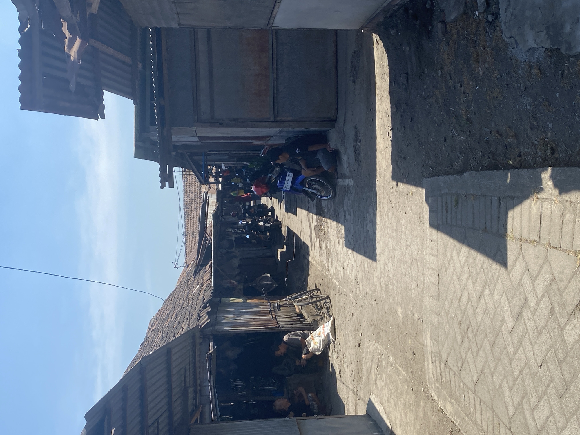 The image depicts a narrow, sun-drenched alleyway in a commercial area, likely a market or a row of small shops, in Kediri City, Indonesia. The buildings lining the alley are rustic, with corrugated metal roofs and walls made of various materials, including corrugated metal and what appears to be concrete blocks. Some structures show signs of wear and tear.
In the foreground, a paved walkway with a herringbone pattern is visible, transitioning into a dirt and gravel path that forms the main thoroughfare of the alley. The alley is lined with parked motorcycles, indicating a common mode of transportation and commerce in the area.
Several individuals are present. On the left side of the alley, in front of one of the shops, two men are seated on the ground. One is wearing a black t-shirt and shorts, and the other a grey t-shirt and shorts. A large, white sack is placed between them. A bicycle is also leaning against the shop.
Further down the alley, on the right side, two men are sitting on the edge of a raised platform in front of a building with a large, metal door. One is wearing a dark t-shirt and shorts, and the other a black t-shirt and grey shorts. A blue motorcycle is parked beside them.
In the background, more motorcycles are visible, suggesting ongoing activity. The sky is clear and blue, with bright sunlight casting strong shadows across the alley. The overall impression is one of a busy, everyday scene in a bustling, traditional urban environment.