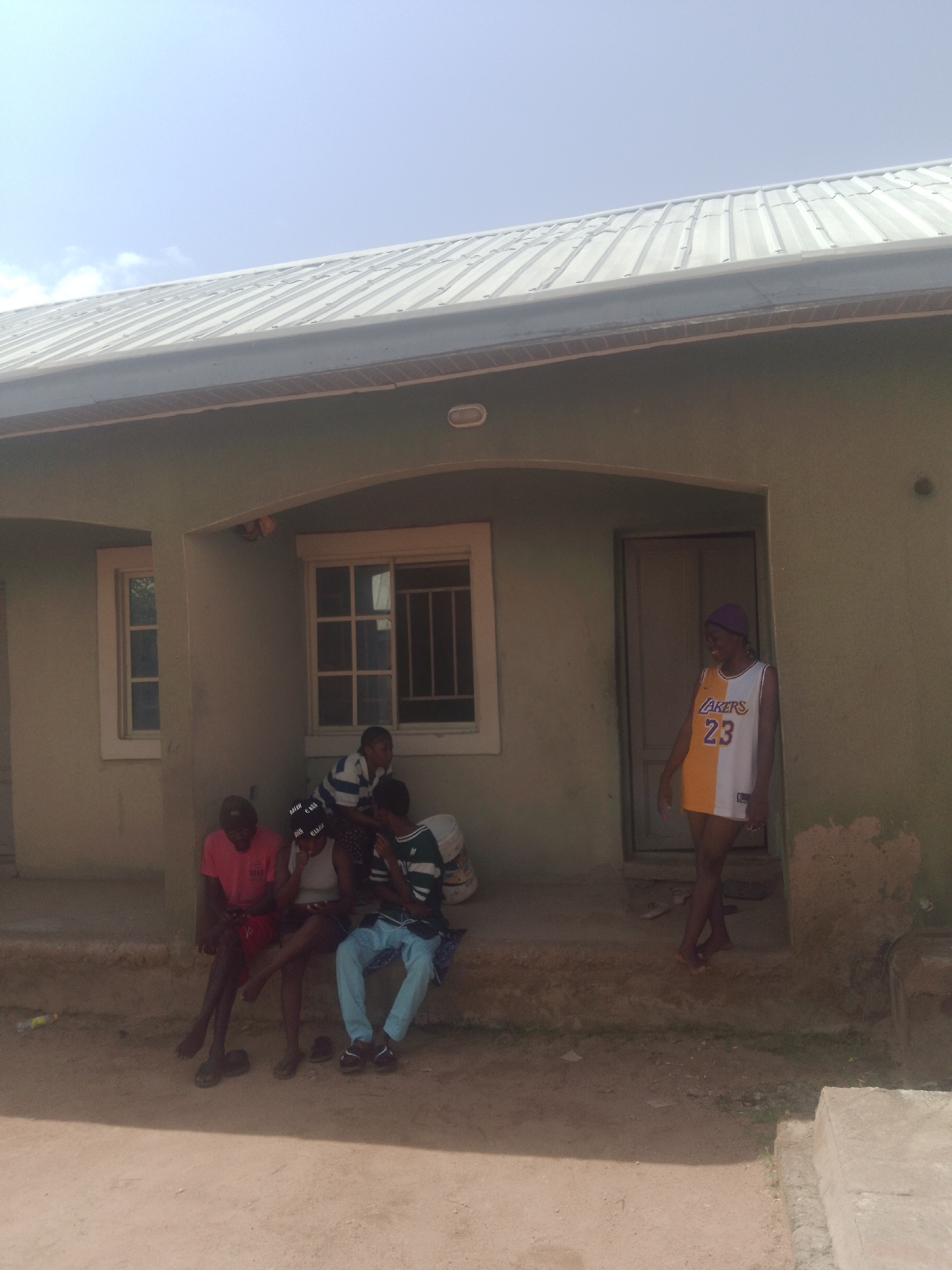 In Jauro Yinu, Nigeria, a group of young people are gathered outside a light green building with a corrugated metal roof. Several individuals are seated on a raised concrete platform in front of the building, while one person stands near the entrance. The people are dressed in casual clothing. The building appears to be a residential structure, with windows and a doorway visible. The ground in front of the building is dirt and sand. The sky above is clear with a hint of blue.