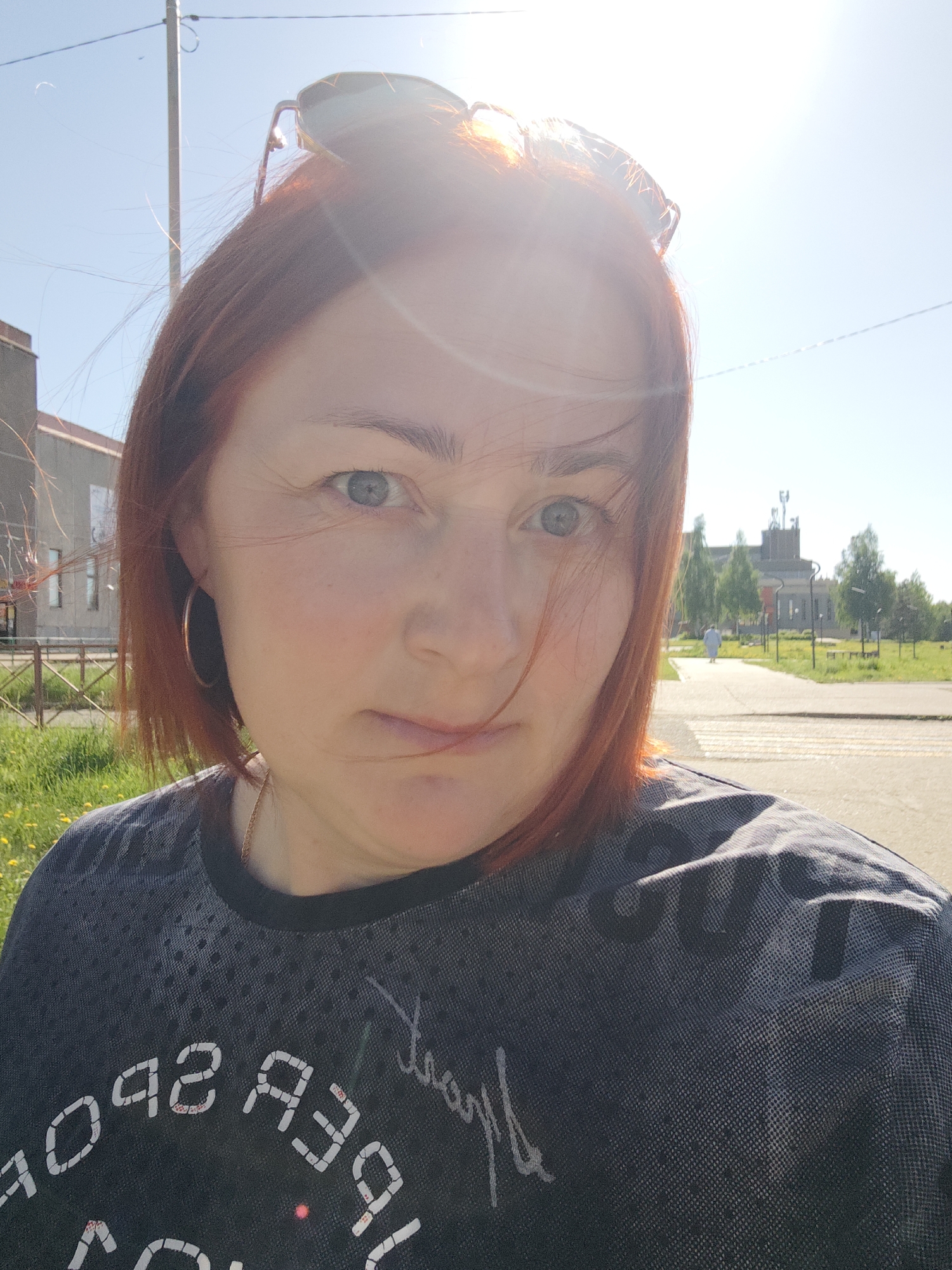A woman with red hair is the primary subject of the photograph. She has fair skin and is wearing a dark t-shirt. Sunglasses are perched on top of her head. The environment includes a street, building, grass, and trees. The location is identified as Марковский, Russia.