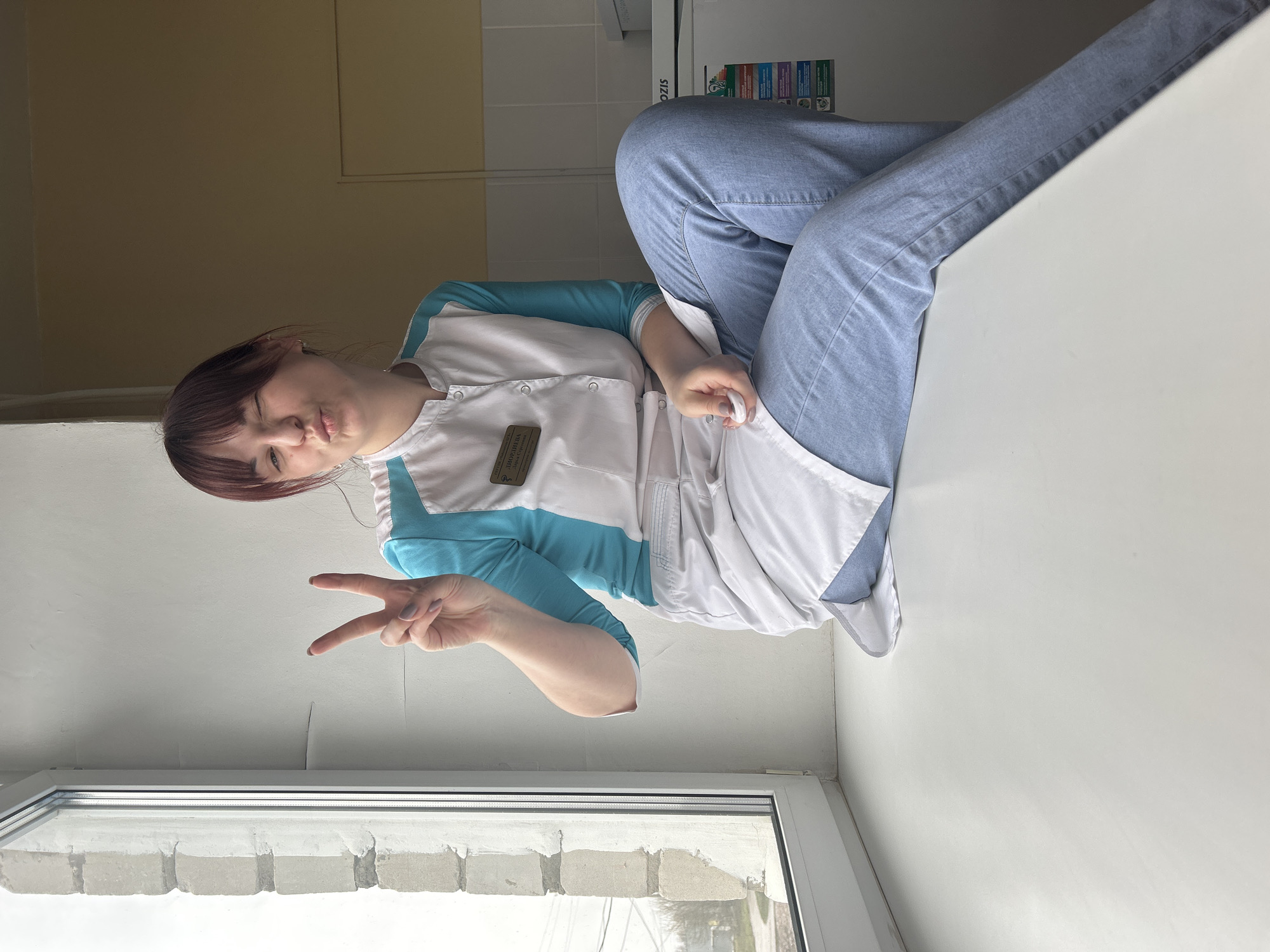 The image shows a woman, likely a medical professional, seated on a windowsill in Фоки, Russia. She's wearing light blue jeans, a white and blue medical scrub top, and white socks. She is making a peace sign with her right hand. Behind her is a tiled wall and a refrigerator with labels. The window is visible in the foreground. The overall lighting is bright and somewhat flat.