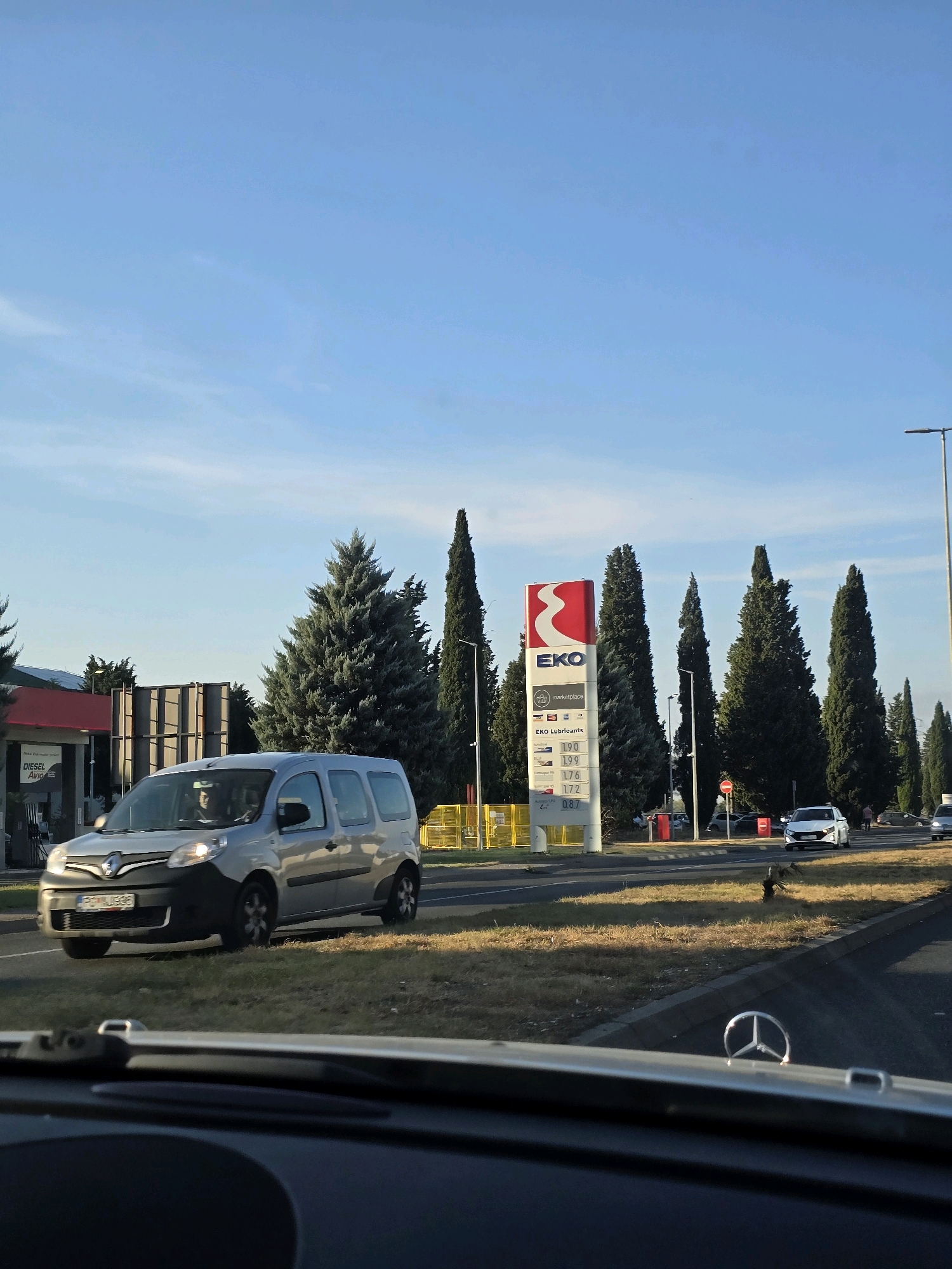 The image depicts a view from inside a vehicle looking out at a gas station and a road on a sunny day in Podgorica, Montenegro. The gas station, identified as EKO, has a prominent red and white sign displaying fuel prices. Various types of fuel are listed, including "LPG" at 0.87. Several cars are visible on the road, with one silver minivan in the foreground and a white car further down the road. Tall cypress trees line the side of the road. The sky is blue with a few wispy clouds. The dashboard of the vehicle is visible in the foreground, including a Mercedes-Benz emblem. The overall scene suggests a typical roadside environment in a city.