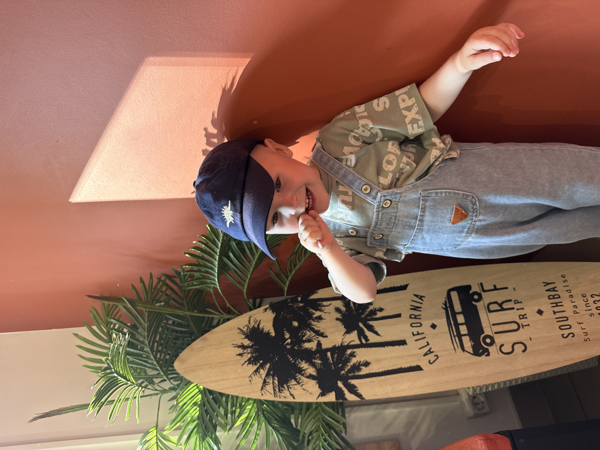 The image shows a young boy wearing a dark blue baseball cap and denim overalls, standing next to a surfboard. The boy is smiling and has his finger in his mouth. The surfboard is decorated with palm trees, a van, and text that reads "CALIFORNIA", "SURF TRIP", and "SOUTHBAY Surf Paradise since 1932". A potted plant with large green leaves is visible to the left of the surfboard. The background is a textured, orange-red wall with a shadow cast by a window or light source. There is no discernible location in Chaikovsky, Russia.