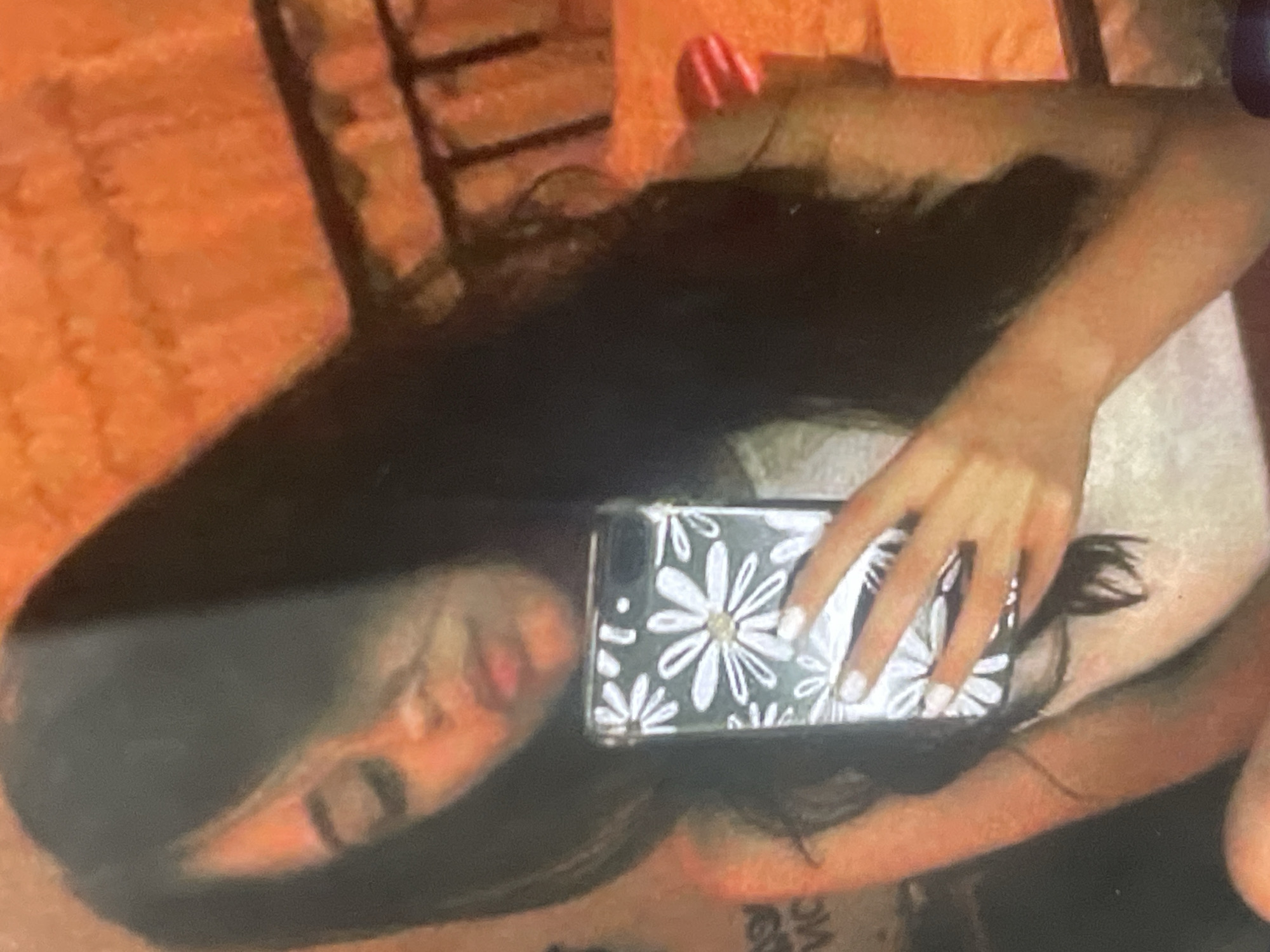This rotated indoor image captures a person, likely female, with long dark hair, lying down while interacting with a smartphone. The main subject is partially obscured by the phone and hair.
The central focus is a hand with light-colored (possibly white) polished fingernails, holding a black smartphone. The phone is encased in a distinctive cover featuring numerous white daisy patterns on a dark background, with a larger, central daisy adorned with a silver or gold-colored center detail. A reflection of the person's face, showing light skin and pinkish lips, is visible on the phone's screen. The person's long, dark hair is spread out around the phone and their head.
The setting appears to be a cozy indoor environment, possibly a bedroom or living area. A light-colored surface, possibly a pillow or blanket, is visible beneath the arm and phone. The background consists primarily of a reddish-orange, textured material, which could be a blanket, rug, or furniture upholstery. Darker elements, possibly parts of a chair or bed frame, are visible in the top corners.
The activity depicted is the person taking a selfie or simply looking at their phone while lying down. Faint, partially obscured text, possibly "VICS" or similar, can be seen in the lower left of the image, and "MC" is barely discernible on the light surface in the lower right, though these are not clearly legible.