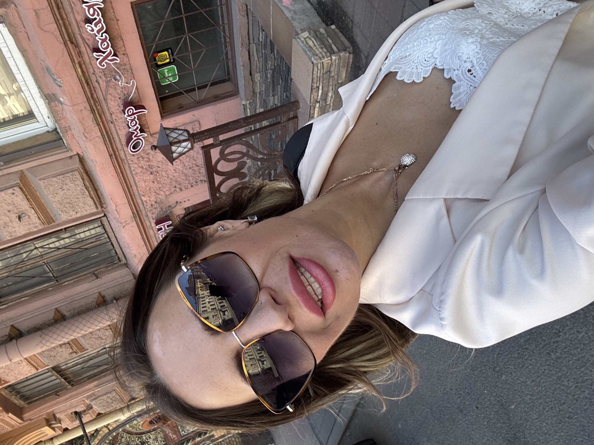 A woman is taking a selfie outdoors in front of a building. The building's facade is pink, with several windows that have metal grates. There is signage on the building that appears to be in Cyrillic script, and one sign reads "Омар Хайям," which translates to "Omar Khayyam," a famous Persian poet. A street lamp is visible to the left of the signage. The woman is wearing sunglasses, a pale pink blazer over a white lace top, and a necklace with a pendant. The reflection in her sunglasses shows buildings with windows. The ground in the foreground is asphalt. The scene suggests an urban environment, possibly a street in Saint Petersburg, Russia, given the architecture and signage.