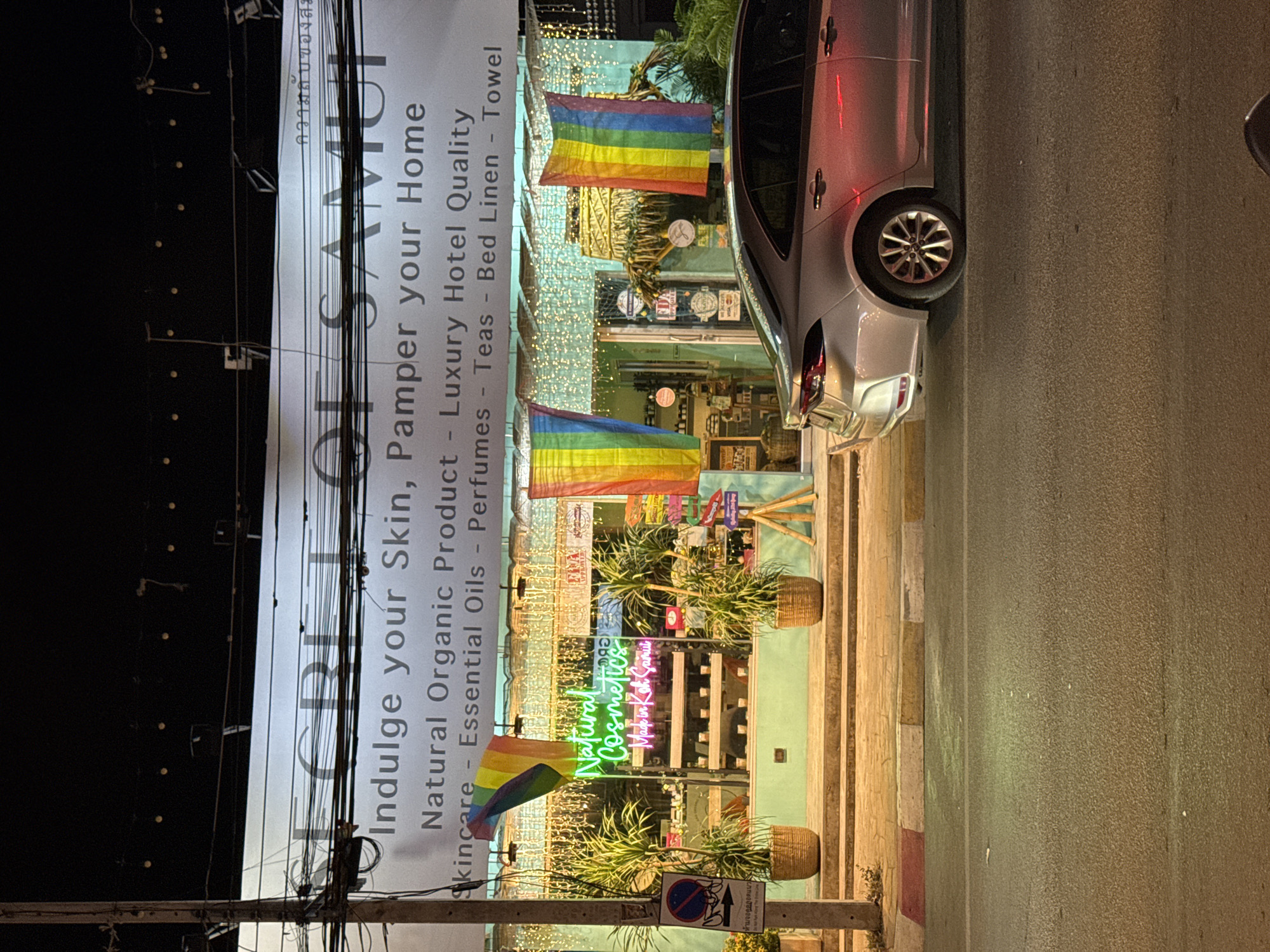 The image depicts a storefront in Baan Bo Phut, Thailand, at night. The storefront, branded "SECRET OF SAMUI," is decorated with rainbow flags and string lights. A neon sign inside reads "Natural Cosmetics Made in Koh Samui." Products are visible on shelves behind the glass. Two potted plants flank the entrance, and a silver sedan is parked on the street in front of the store. A traffic sign is visible on a pole to the left of the storefront. The street in front of the building has a sidewalk with yellow and red markings. The overall scene suggests a commercial establishment operating in a coastal town.