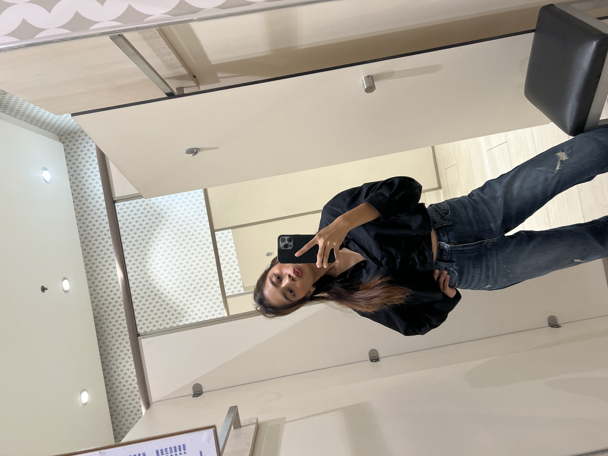 A young woman is depicted taking a mirror selfie inside a fitting room, likely within a retail store or department store in Davao City, Philippines. She stands centrally in the frame, holding a black smartphone with multiple rear cameras in her left hand to capture her reflection, while her right hand is placed on her hip. She has long, dark brown hair and is wearing a black collared, button-down shirt paired with dark blue denim jeans that feature distressed areas. Her facial expression is a 'pout' or 'kissing face' directed towards the phone.
The fitting room features light-colored, possibly off-white or beige, partition walls. A silver hook is mounted on the left wall of the cubicle. The reflected environment reveals a white ceiling with two circular recessed light fixtures and a small, dark object, possibly a sprinkler head, centrally suspended. Behind the cubicle, a wall section with a light grey and white geometric diamond pattern is visible, along with a horizontal metal hanging rail. To the far left in the reflection, a white sign displaying prices in Philippine Pesos (e.g., "P 390," "P 265," "P 450") is partially visible. The floor is covered with light-colored tiles. A black, rectangular stool with a silver metal frame is positioned in the lower right corner of the reflected scene.