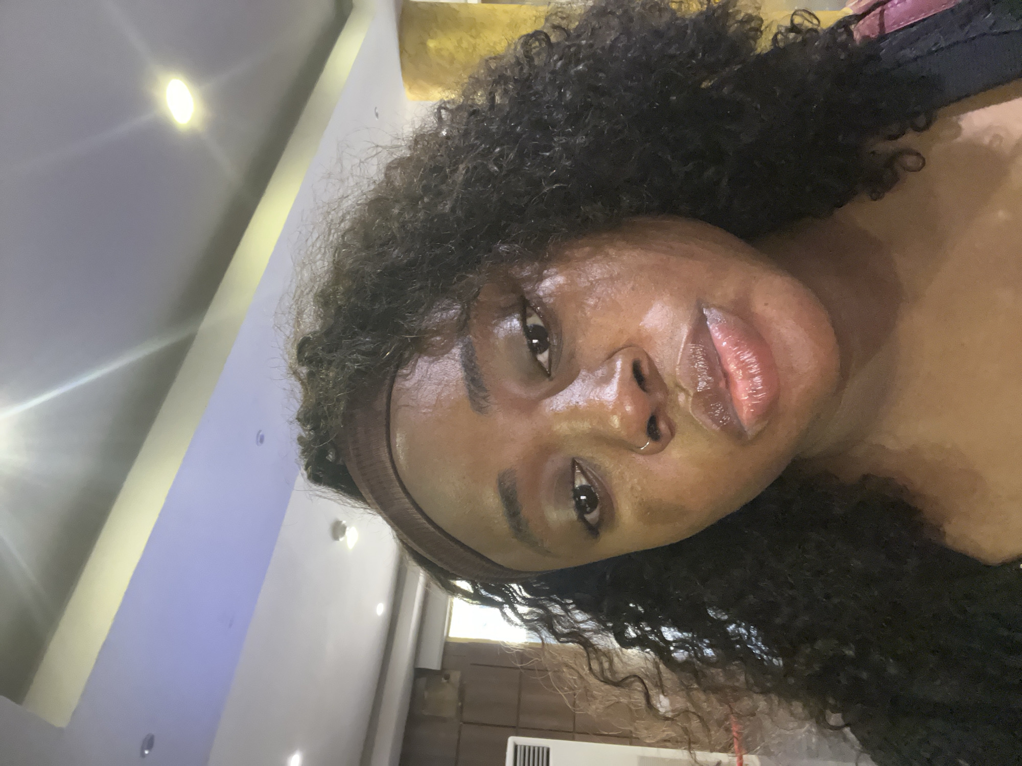 A close-up, slightly angled, front-facing selfie of a young Black woman indoors. She has curly, dark brown hair that is pulled back by a brown headband. Her skin appears to be slightly dewy or oily, and she is wearing subtle makeup, with a hint of lip gloss. Her eyes are dark brown, and she has a small hoop nose ring. The background suggests an indoor setting with a ceiling that features recessed lighting and a yellow accent strip. To the left, a white air conditioning unit is partially visible. The overall lighting is bright, with some glare from the ceiling lights. The woman's expression is neutral to slightly pensive. The location is identified as Abuja, Nigeria.