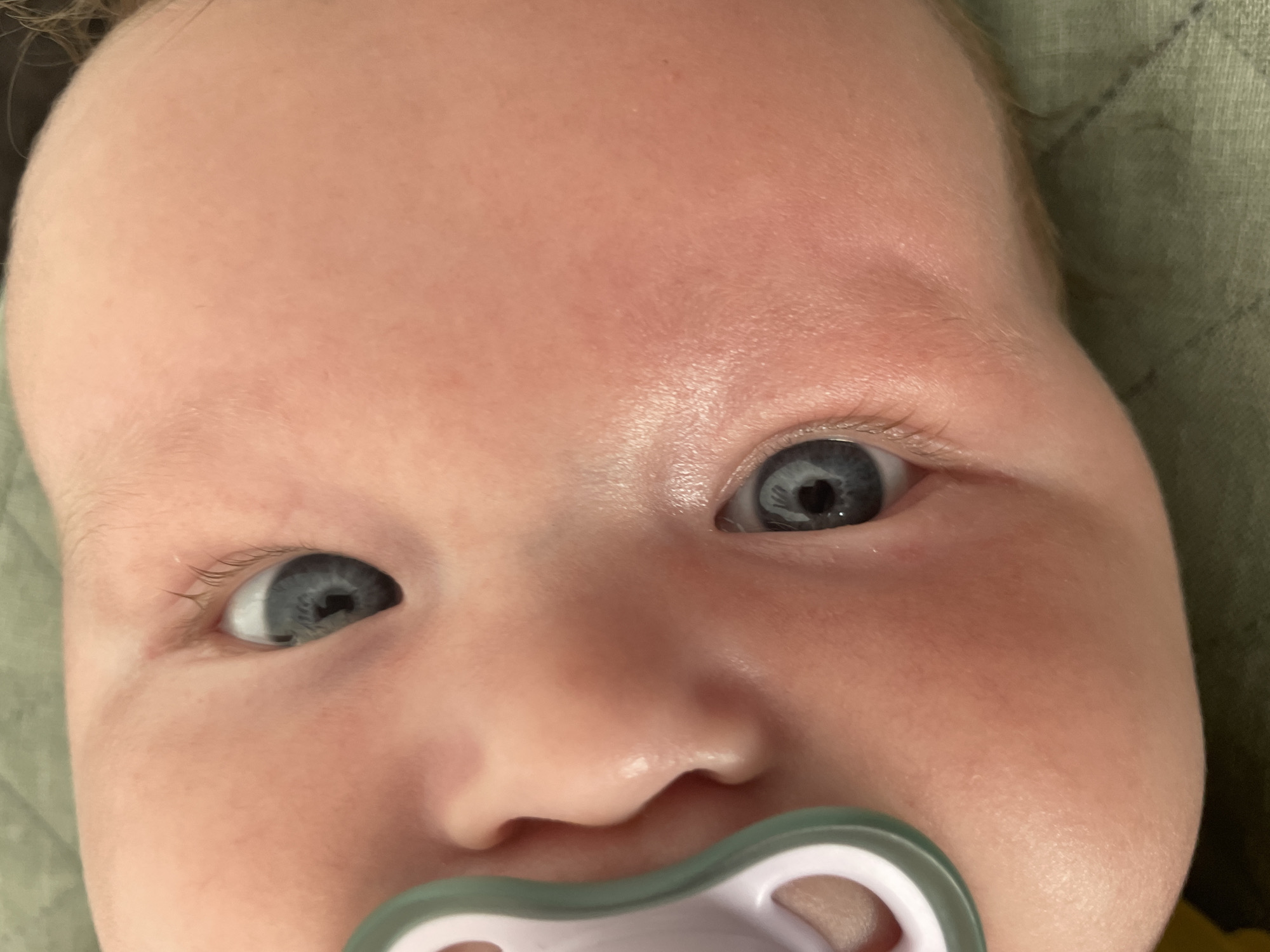The image is a close-up, head-on, Dutch angle shot of a baby's face, tilted to the left. The baby has light skin, noticeable rosy cheeks, and light eyelashes. The left eye is fully visible, with a blue iris and a dark pupil. The right eye is partially visible and also shows a blue iris. A light pink pacifier with a light teal rim is positioned to the left of the baby's face, partially obscuring the left cheek and mouth area. The background consists of a textured, muted green fabric. The image is oriented such that the baby's head is positioned towards the top right of the frame. The overall lighting is soft and even, highlighting the baby's skin texture. The location in Stavropol, Russia is not discernible from the image.