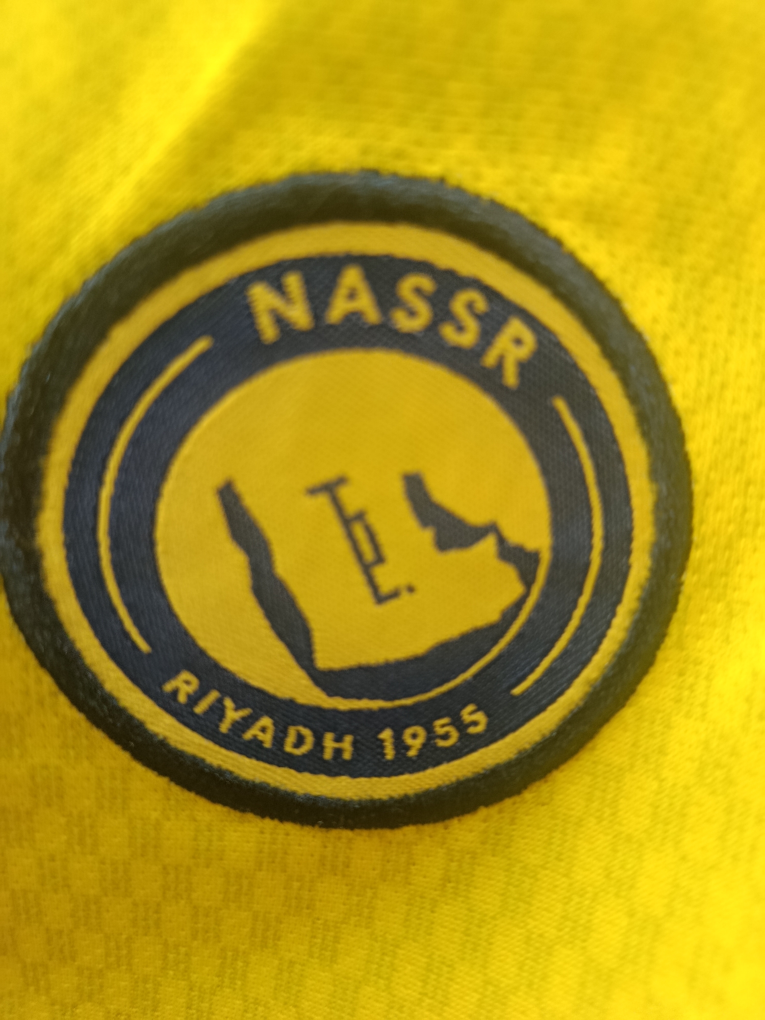 The image shows a close-up of a yellow fabric with a circular emblem on it. The emblem has a dark blue border and a yellow inner circle. Inside the inner circle, there is a silhouette of what appears to be the Arabian Peninsula in dark blue, with some Arabic script in dark blue on the left side. The word "NASSR" is written in large, dark blue capital letters in an arc above the silhouette. Below the silhouette, the words "RIYADH 1955" are written in yellow capital letters, also in an arc. The fabric appears to be a jersey or shirt. The emblem is likely a sports team logo. There is no indication of the location being Sertolovo, Russia.