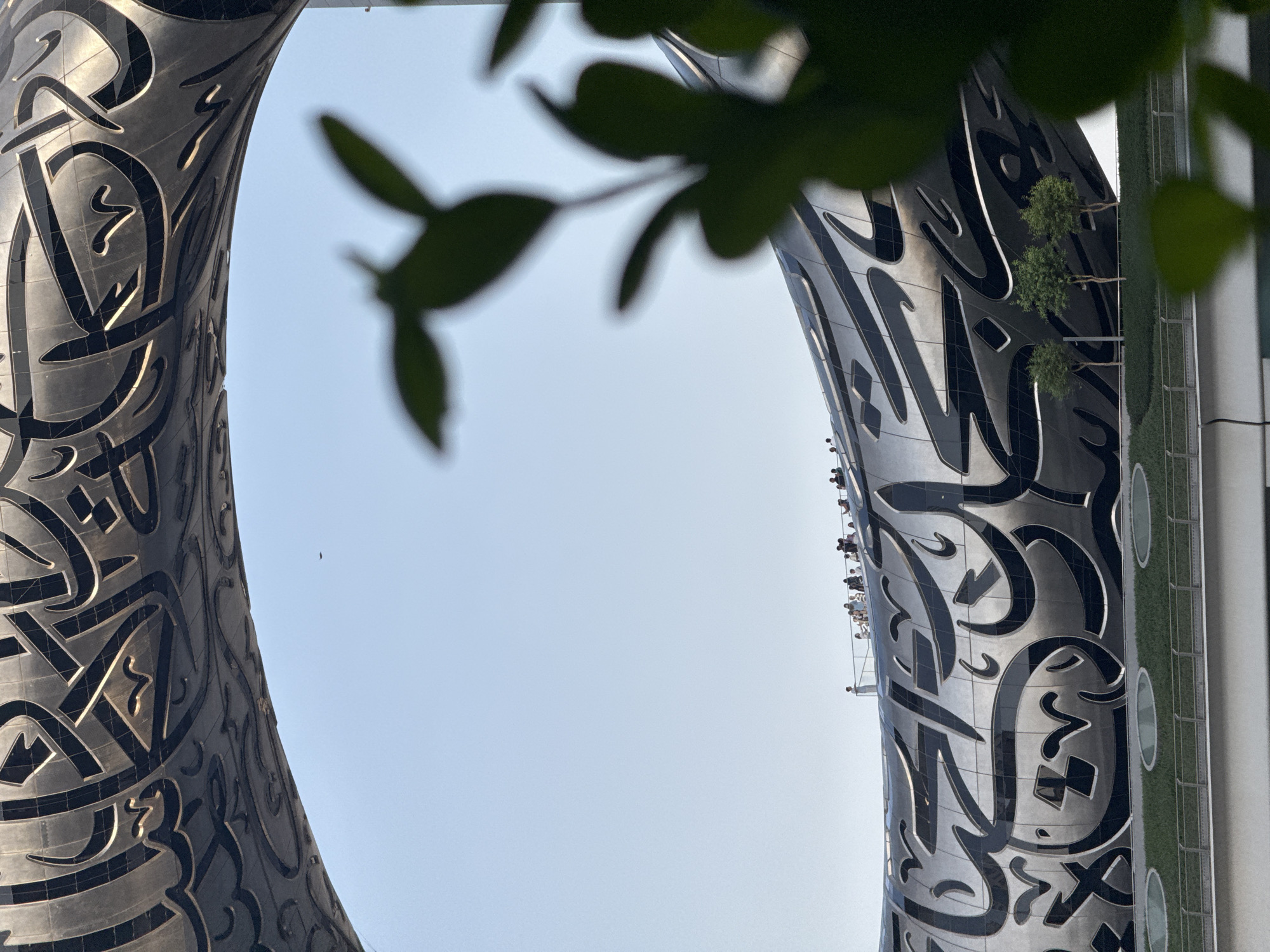 The image shows the Museum of the Future in Dubai, United Arab Emirates. The building's distinctive torus-shaped facade is decorated with Arabic calligraphy. In the foreground, out-of-focus green leaves frame the scene. In the middle ground, a group of people stand on a balcony overlooking the cityscape. The background consists of a clear blue sky, with a small black dot that might be a bird or an aircraft. The lower part of the image shows the green landscaping around the museum, with some trees.