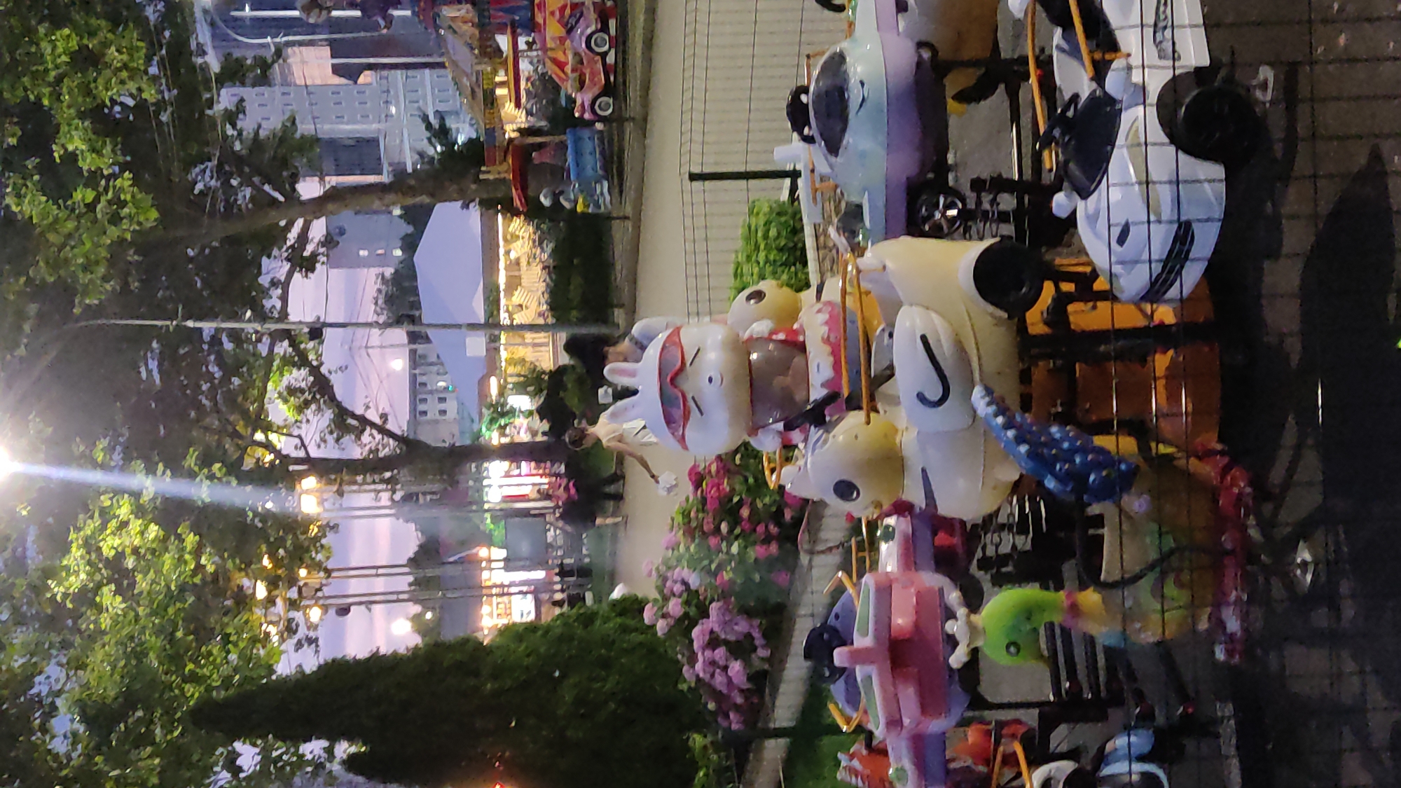 The image depicts a scene at an amusement park in Krasnodar, Russia. The foreground features a carousel with various colorful rides, including a white rabbit character wearing sunglasses and a brown base, duck-shaped seats, a green bird-like ride with a crown, and white car-shaped rides. The carousel is surrounded by a black metal fence. In the middle ground, there are people walking and some amusement park stalls with tents and lights. The background reveals tall buildings and trees, illuminated by streetlights and the twilight sky. The overall atmosphere suggests an evening recreational setting.