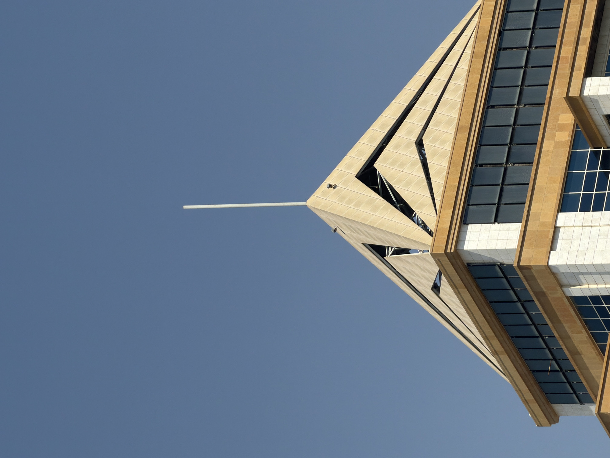 A close-up, low-angle shot captures the apex of a modern building against a clear, bright blue sky. The building's roof features a distinctive pyramidal design with stepped, beige-toned panels. A thin, white antenna rod extends vertically from the very top of the pyramid. Below the apex, a section of the building is visible, clad in white stone and accented with a band of dark-tinted windows. A horizontal beige trim runs along the top of this section. The architectural style suggests a contemporary urban setting. There are no people or animals in the frame, and no discernible activity. The lighting indicates it is daytime, likely a sunny day, with soft shadows cast on the building's surfaces.