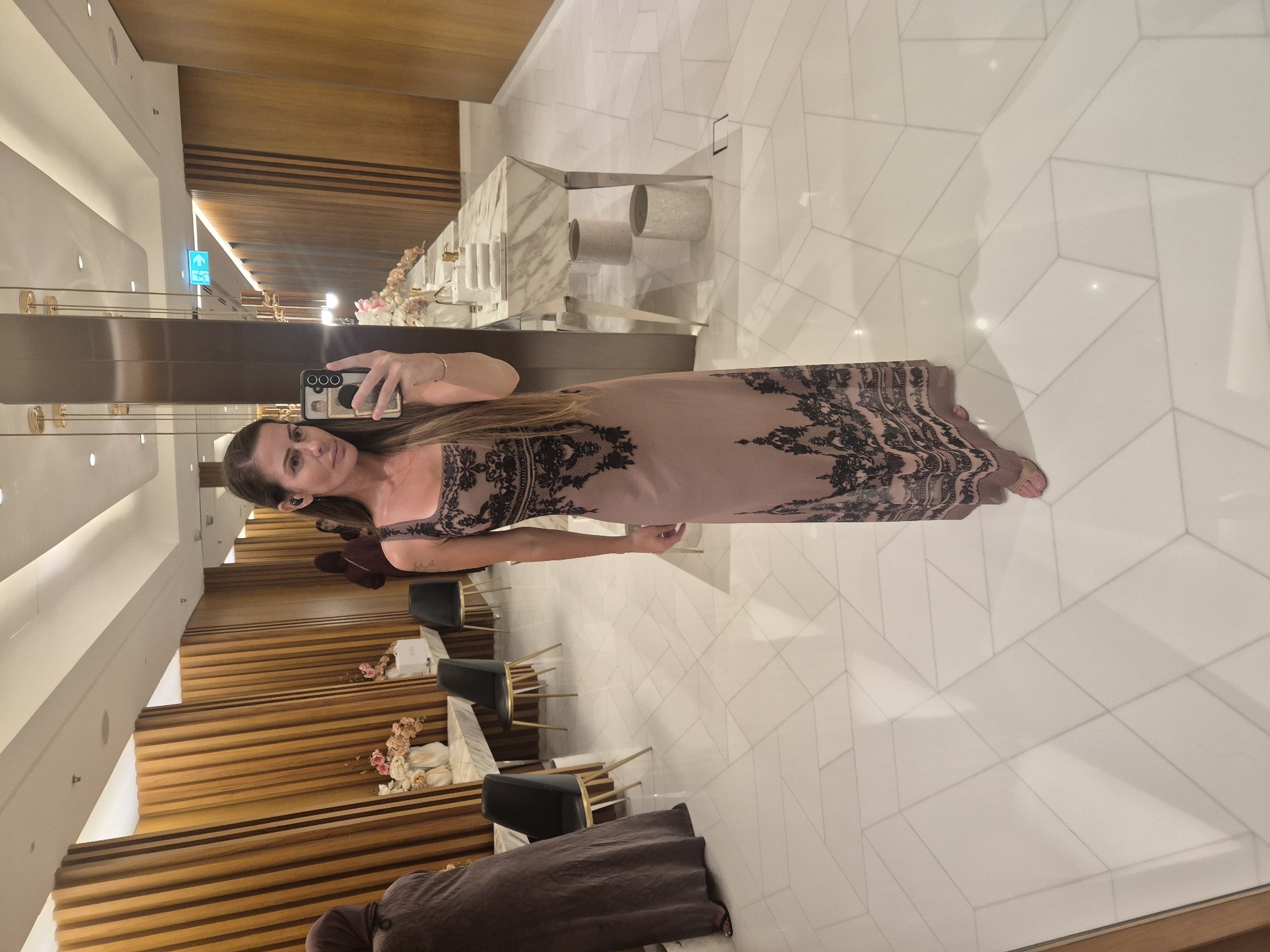 The image, oriented on its side, captures a woman taking a mirror selfie in what appears to be a modern, upscale indoor setting, possibly a hotel lobby, hallway, or a restaurant/lounge.
The primary subject is a woman, seen from the waist up in the mirror's reflection. She has long, dark brown hair, parted on the side, and is looking directly into the camera. She wears a long, light brown or dusty pink sleeveless dress adorned with intricate black, lace-like patterns that extend down the sides and across the bodice. Her expression is neutral to slightly smiling. She holds a dark-colored smartphone with her right hand, positioned in front of her face. The phone's screen displays what looks like a wallpaper or another image, with some visible icons indicating a camera interface. She has a faint tattoo on her left forearm.
The background, also reflected in the mirror, showcases a sophisticated interior design. To the woman's left, a section of a richly textured wooden slatted wall is visible, reflecting the warm lighting. Above this, a green exit sign with a white arrow points to the left. To her right, a polished white marble counter or table is featured, holding several decorative vases or bowls, some containing light-colored floral arrangements. Behind these items, a white tiled wall with a geometric pattern is visible. The floor is covered in large, light-colored, possibly off-white, geometric tiles that create a subtle, interlocking pattern. The woman's feet are visible at the bottom of the frame, wearing pink open-toed shoes. Further into the reflected background, several chairs with black seats and golden frames are arranged around tables, suggesting a seating area. The overall impression is one of elegance and contemporary style, illuminated by artificial lighting, indicating an indoor setting, likely in the evening or night.