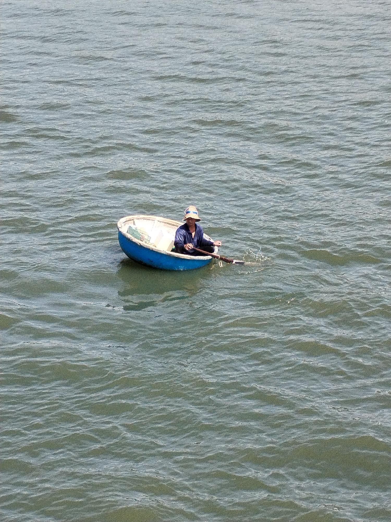A lone individual in a wide-brimmed hat and dark blue clothing is seated in a small, round, blue boat. The boat is on calm, greyish-blue water with gentle waves. The person is holding a long, thin pole, likely a fishing rod or oar, extending out to the right side of the boat, where a small splash is visible in the water. The boat appears to be made of fiberglass or a similar material, with a white interior. The scene is set in an open body of water, suggesting a marine or large lake environment. The overall impression is one of quiet activity on a serene day.